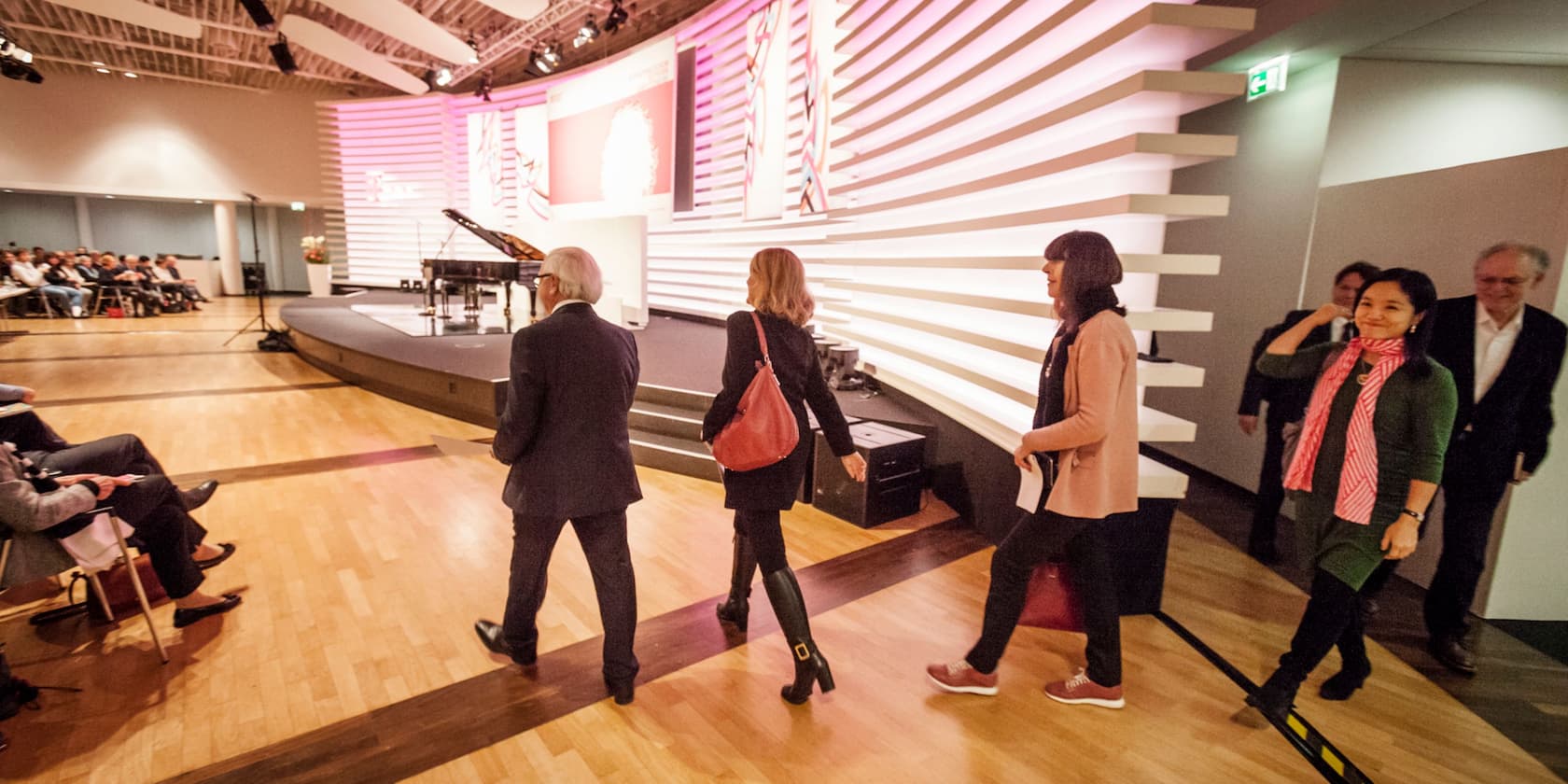 A group of people leaving a stage at a conference or event. The audience is seated on the left side.