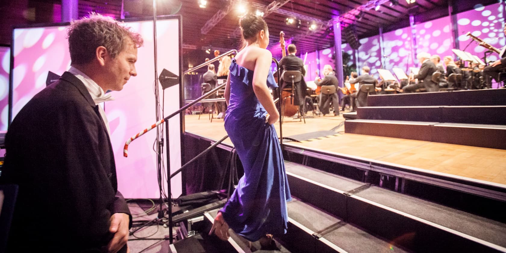 A woman in a blue dress and a man in a suit are entering a stage where an orchestra is performing.