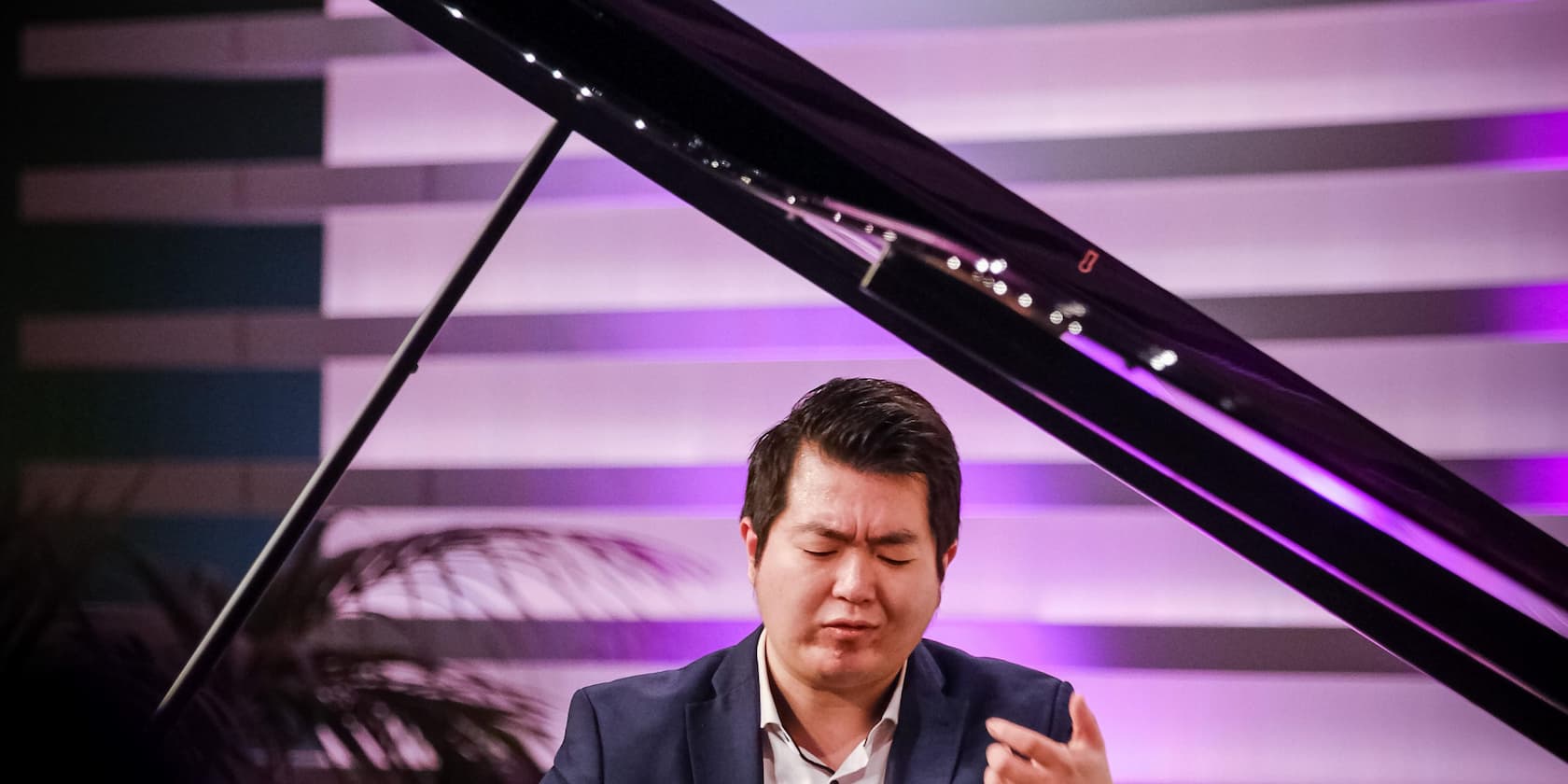 A man plays the piano passionately with closed eyes in front of a purple-lit background.