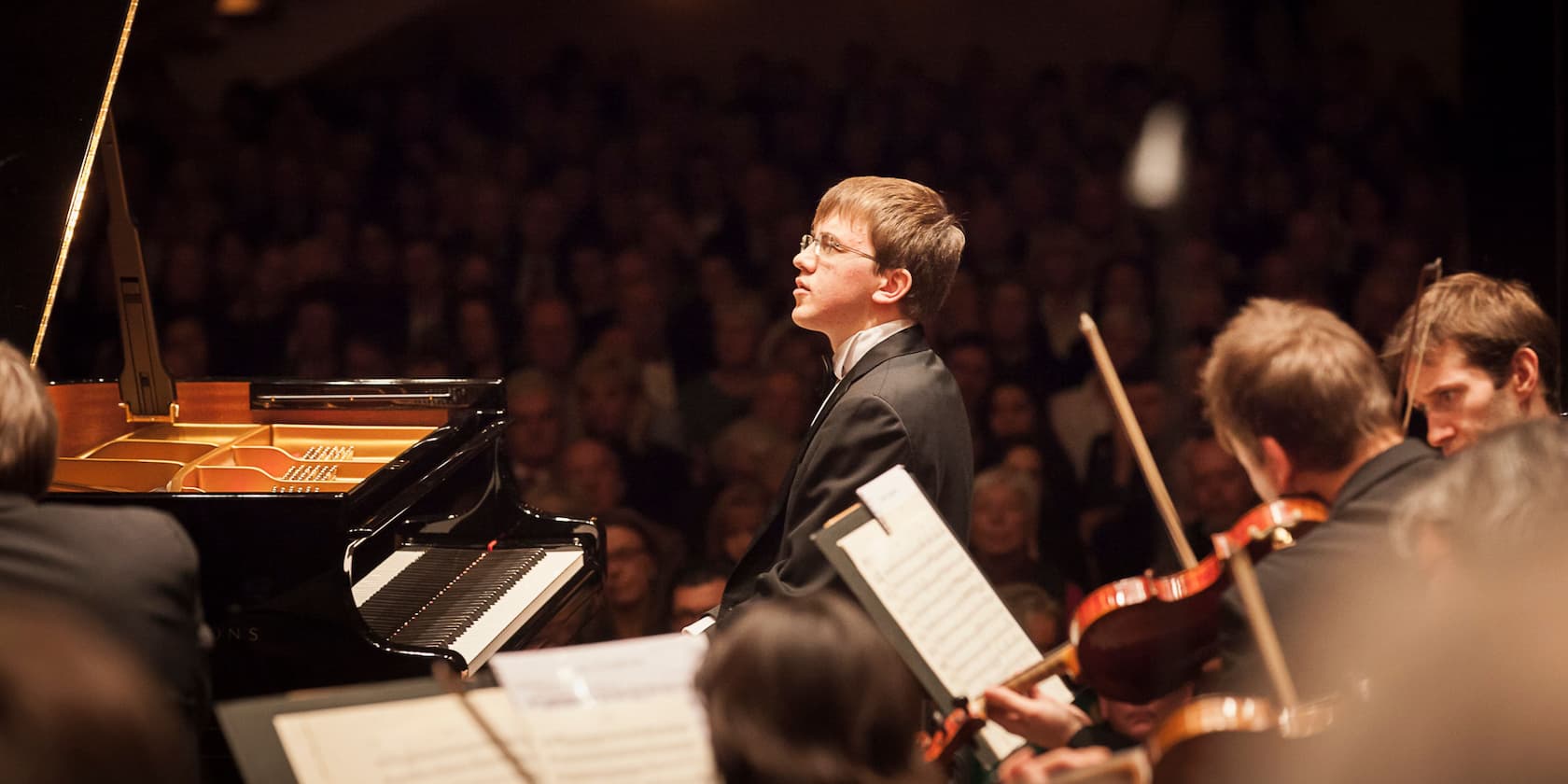 Ein junger Musiker spielt Klavier bei einem Konzert, begleitet von einem Orchester im Hintergrund.