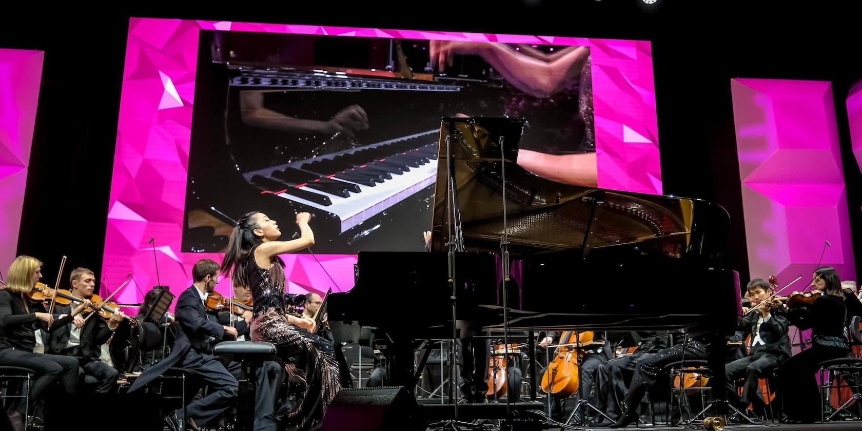 A pianist performs on a grand piano, surrounded by an orchestra. A large screen in the background shows a close-up of hands on the keys. The background is pink.