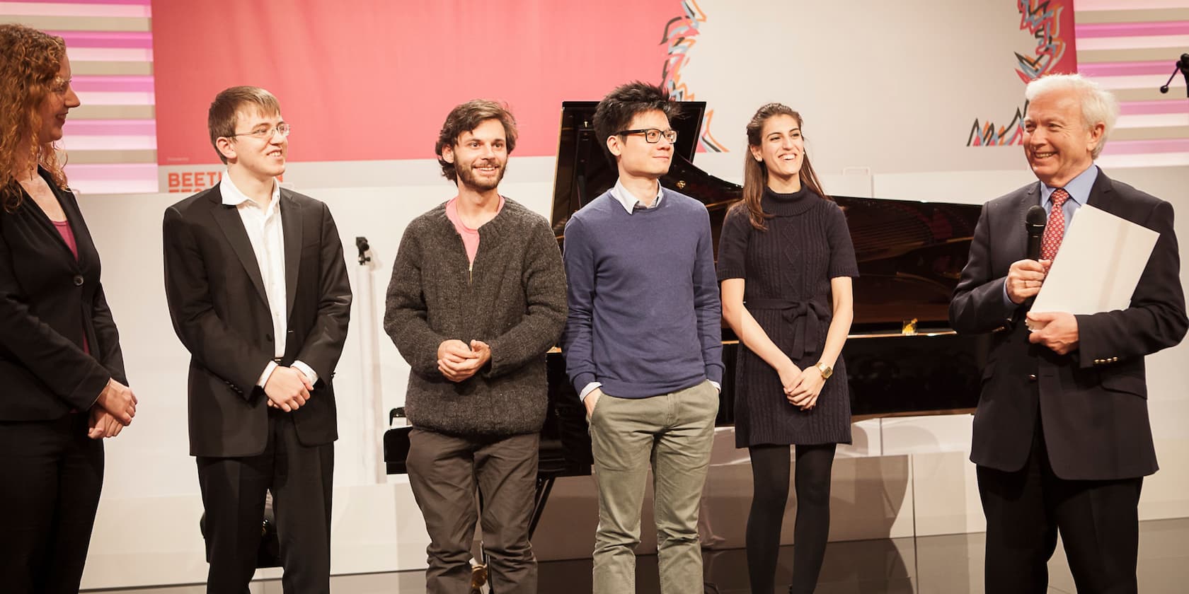 Five people stand side by side on stage, one person holding a microphone, likely during an event or ceremony.