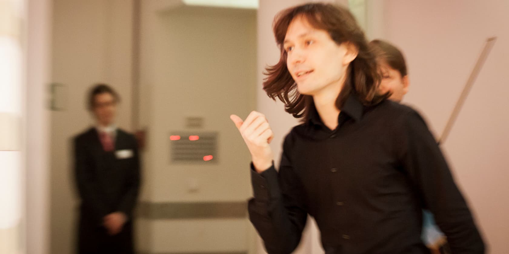 A person in a black shirt gesturing in a blurry room, with a second person standing in the background.