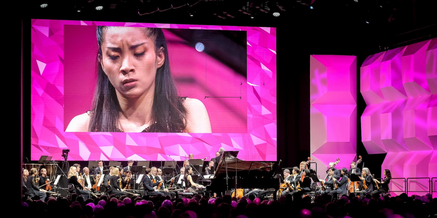 A pianist performs with an orchestra on stage. In the background, a large display shows a close-up of the pianist.