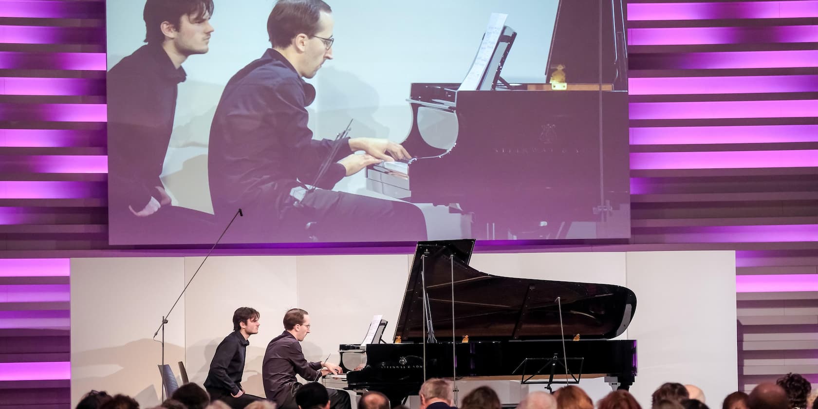 Two musicians are playing a grand piano on a stage while the audience watches. A large projection of the performance is visible in the background.