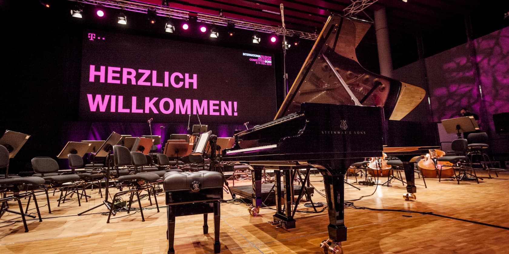 A concert hall with empty chairs and a grand piano. On stage, there is a large sign with the text 'HERZLICH WILLKOMMEN!' in pink neon lights.