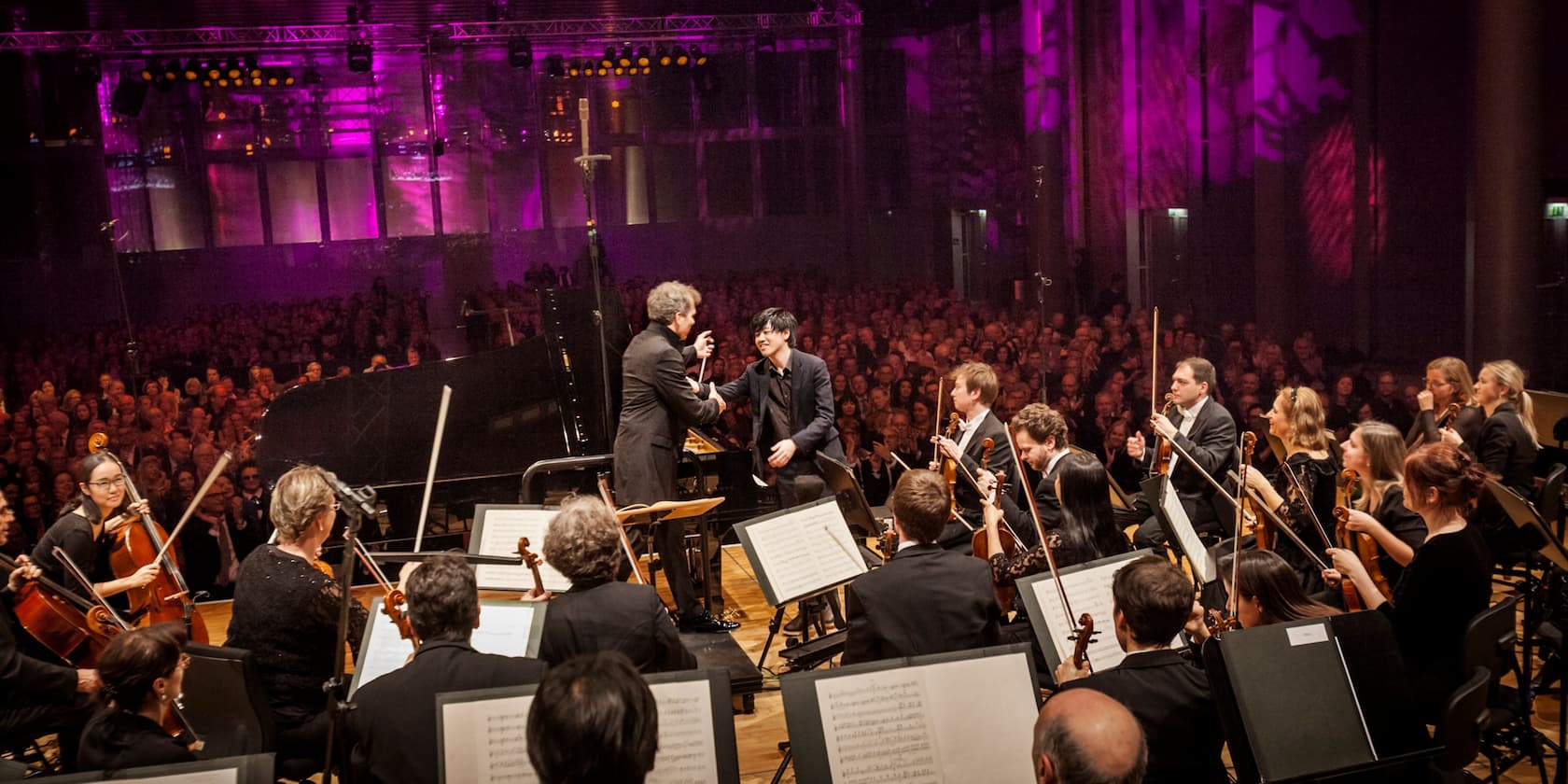 A conductor and a pianist bowing and shaking hands after a concert performance in front of a large audience in a concert hall with purple lighting.