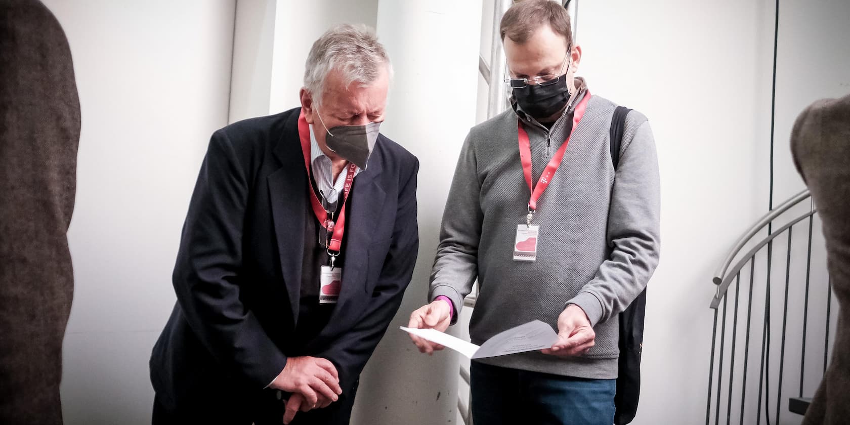 Two men wearing masks and red ID badges reading documents together.