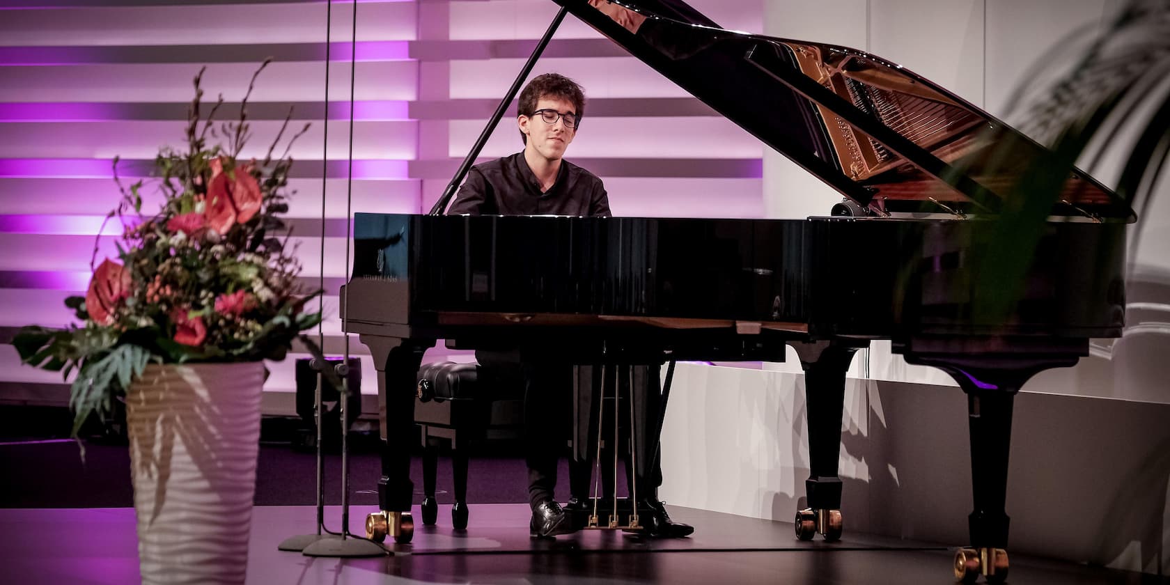 Man playing piano on a stage next to a flower arrangement.