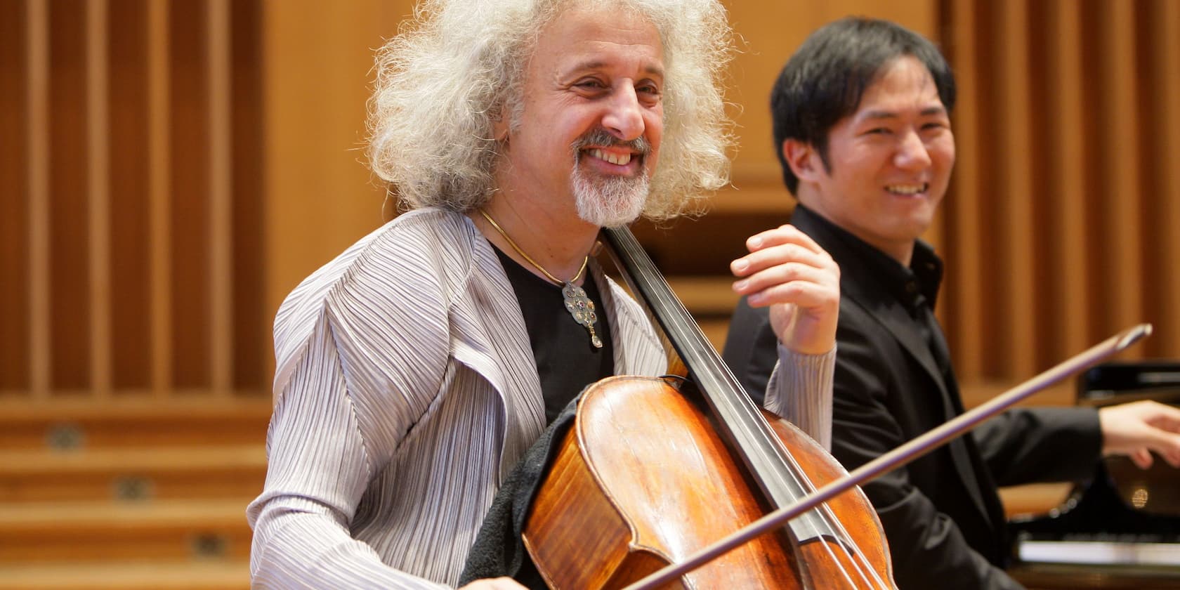 Two musicians, one playing the cello and the other playing the piano, laughing during a performance.