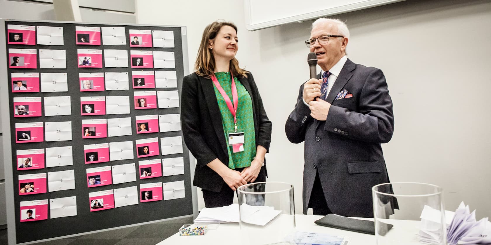 A man holds a microphone and speaks while a woman stands next to him in front of a bulletin board filled with photos.