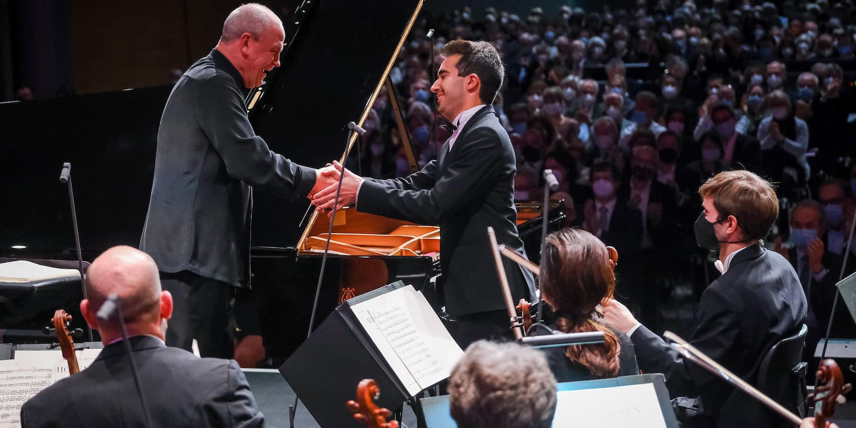 A conductor and pianist shake hands during a concert in front of an applauding audience.