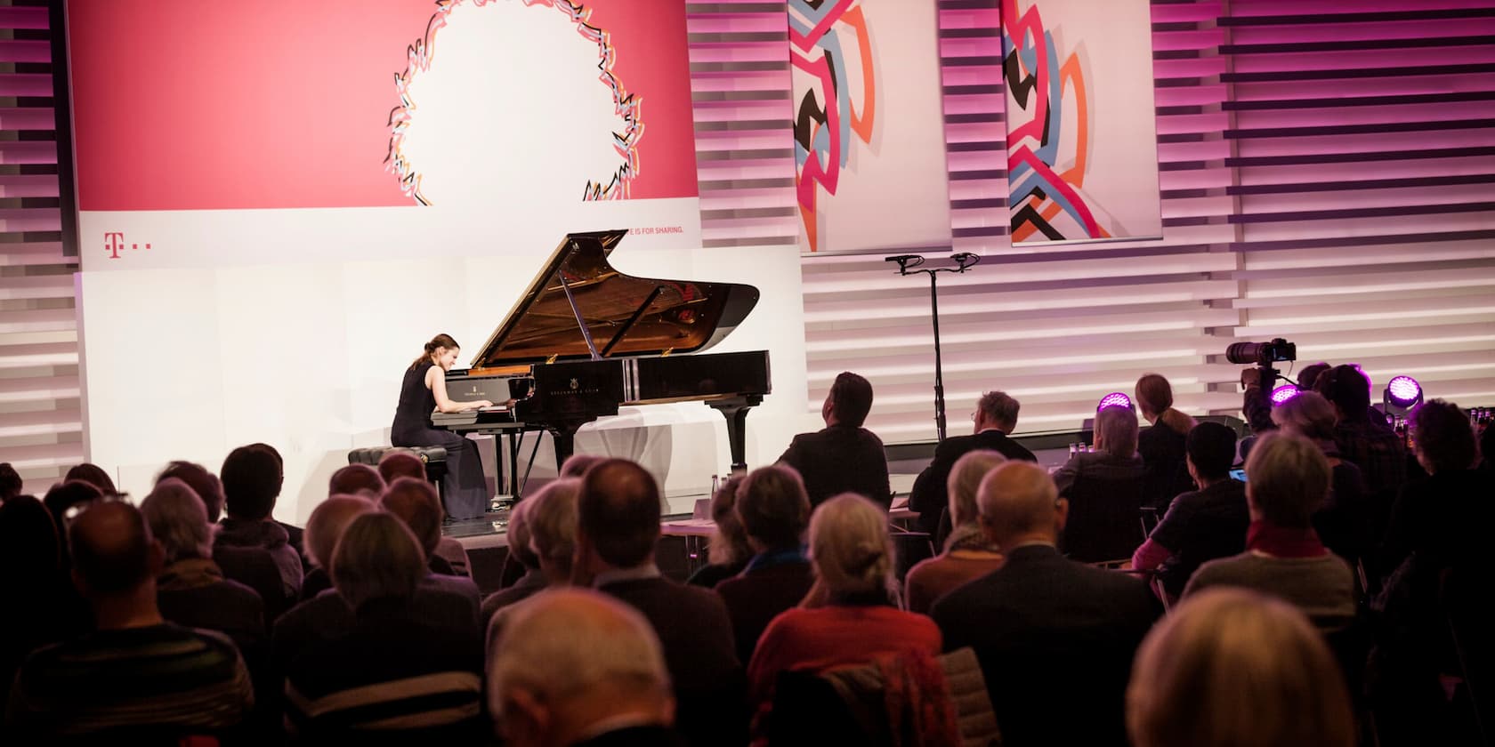 A pianist performs on stage in front of an audience. In the background, there is a large advertisement banner for Deutsche Telekom.