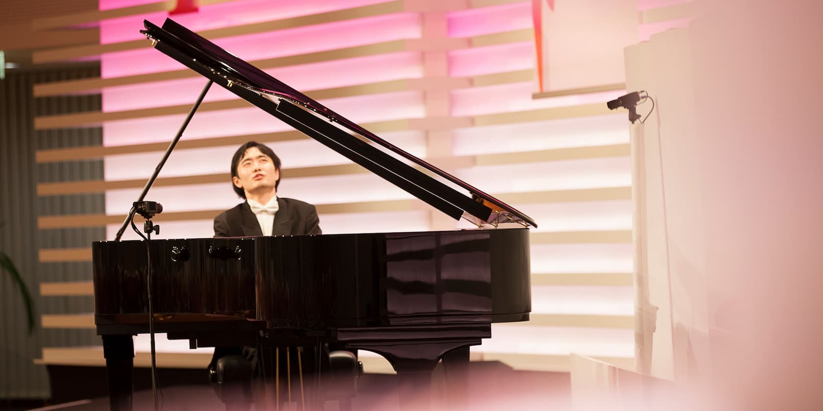 A man playing piano in a concert hall with pink and white lights in the background.