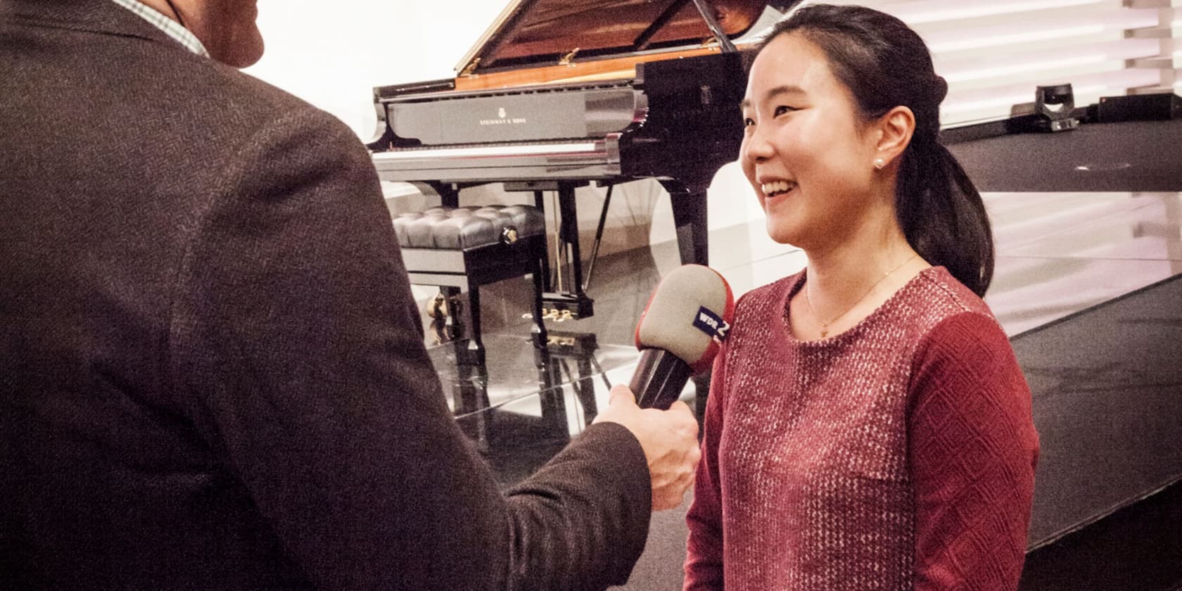 A woman is being interviewed by a reporter holding a microphone. There is a piano in the background.