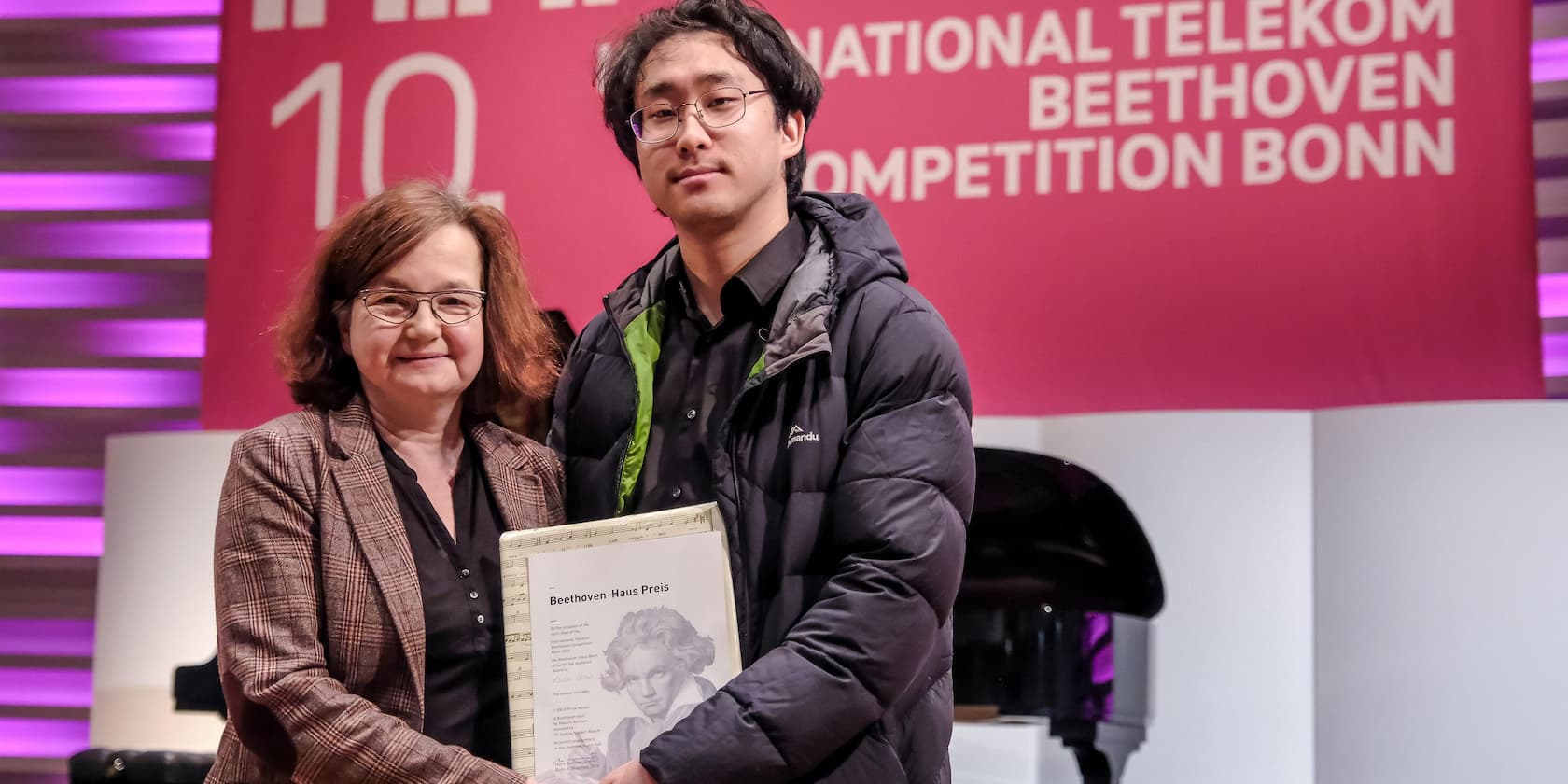 Zwei Personen stehen auf der Bühne bei der International Telekom Beethoven Competition Bonn; eine Person hält eine Urkunde mit der Aufschrift 'Beethoven-Haus Preis'.