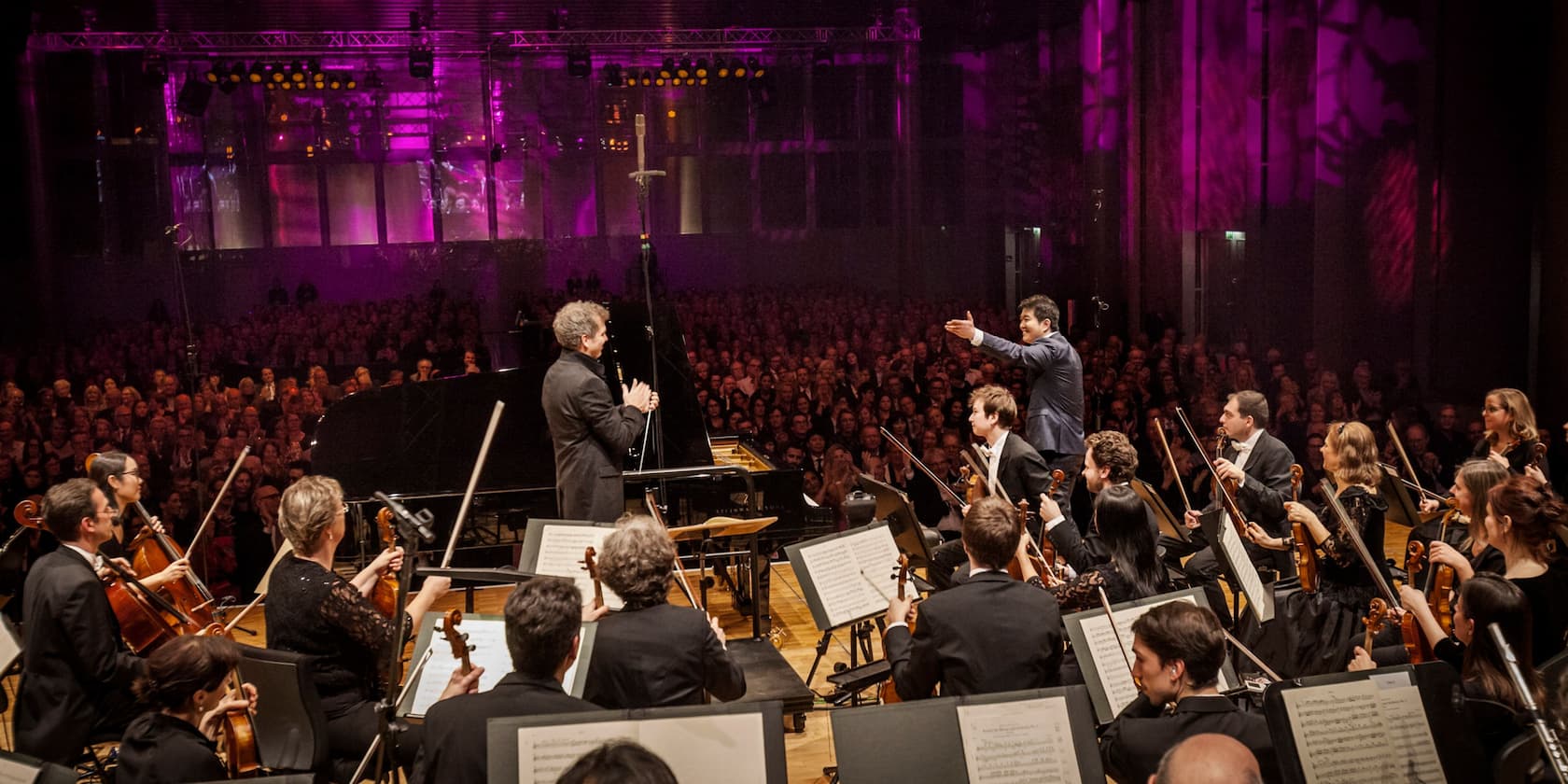 An orchestra performs in front of a large audience in an atmospherically lit hall.