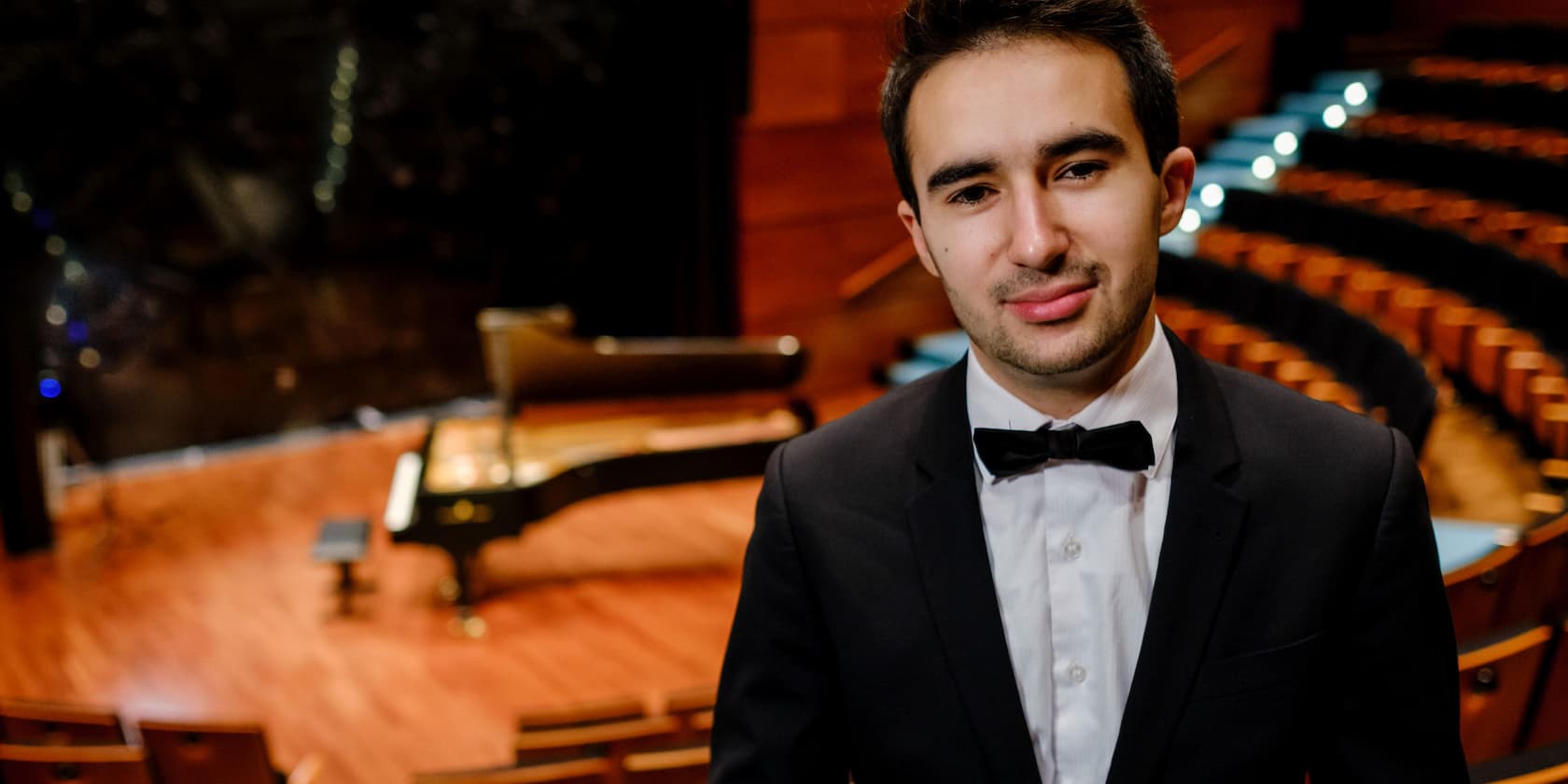 A young man in a suit and bow tie stands on an empty stage with a grand piano in the background.