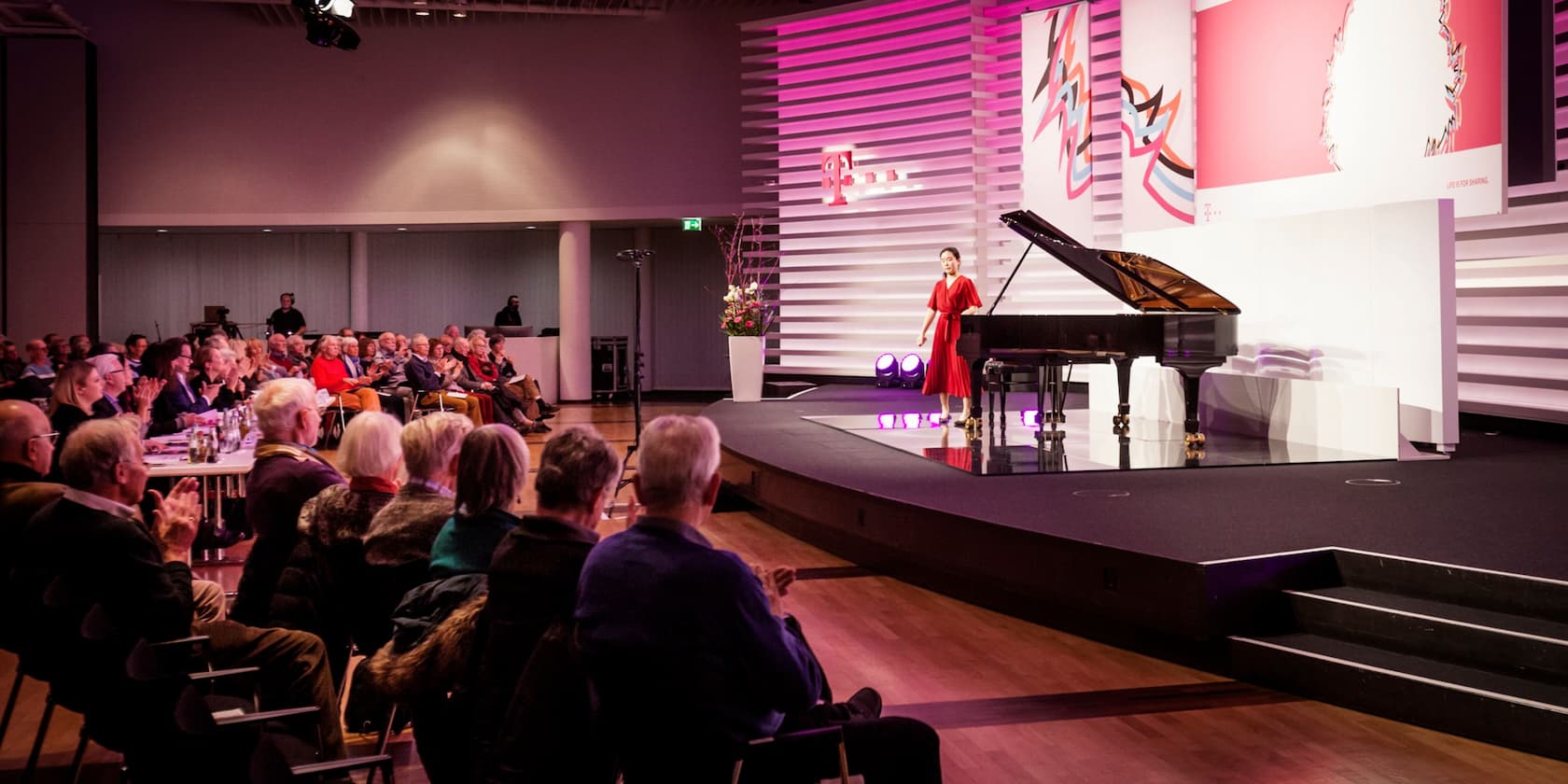 A woman in a red dress stands beside a grand piano on a stage while an audience applauds.