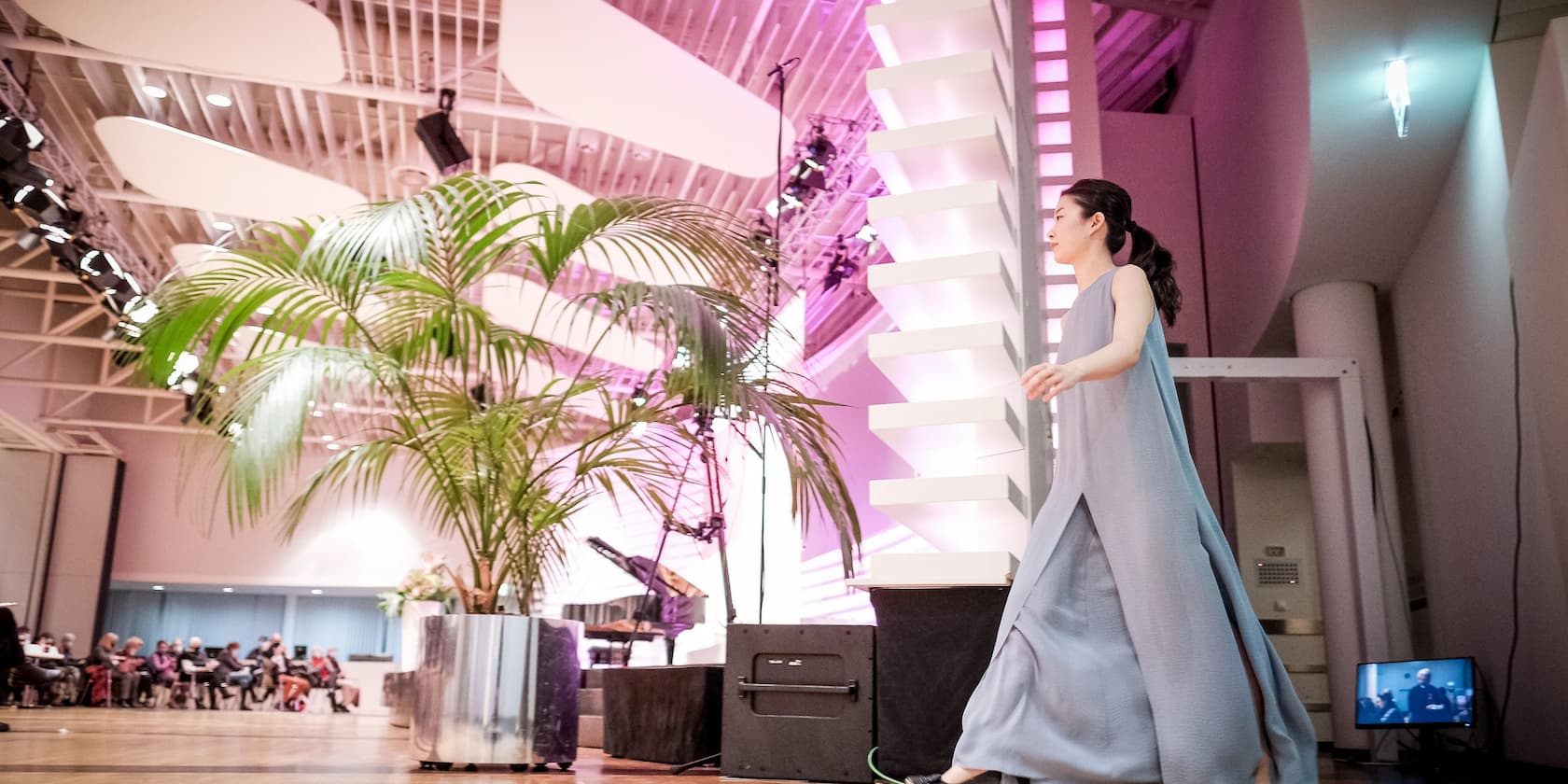 A woman in a blue dress walks along a stage at an event. In the background, there are audience members and a large houseplant.