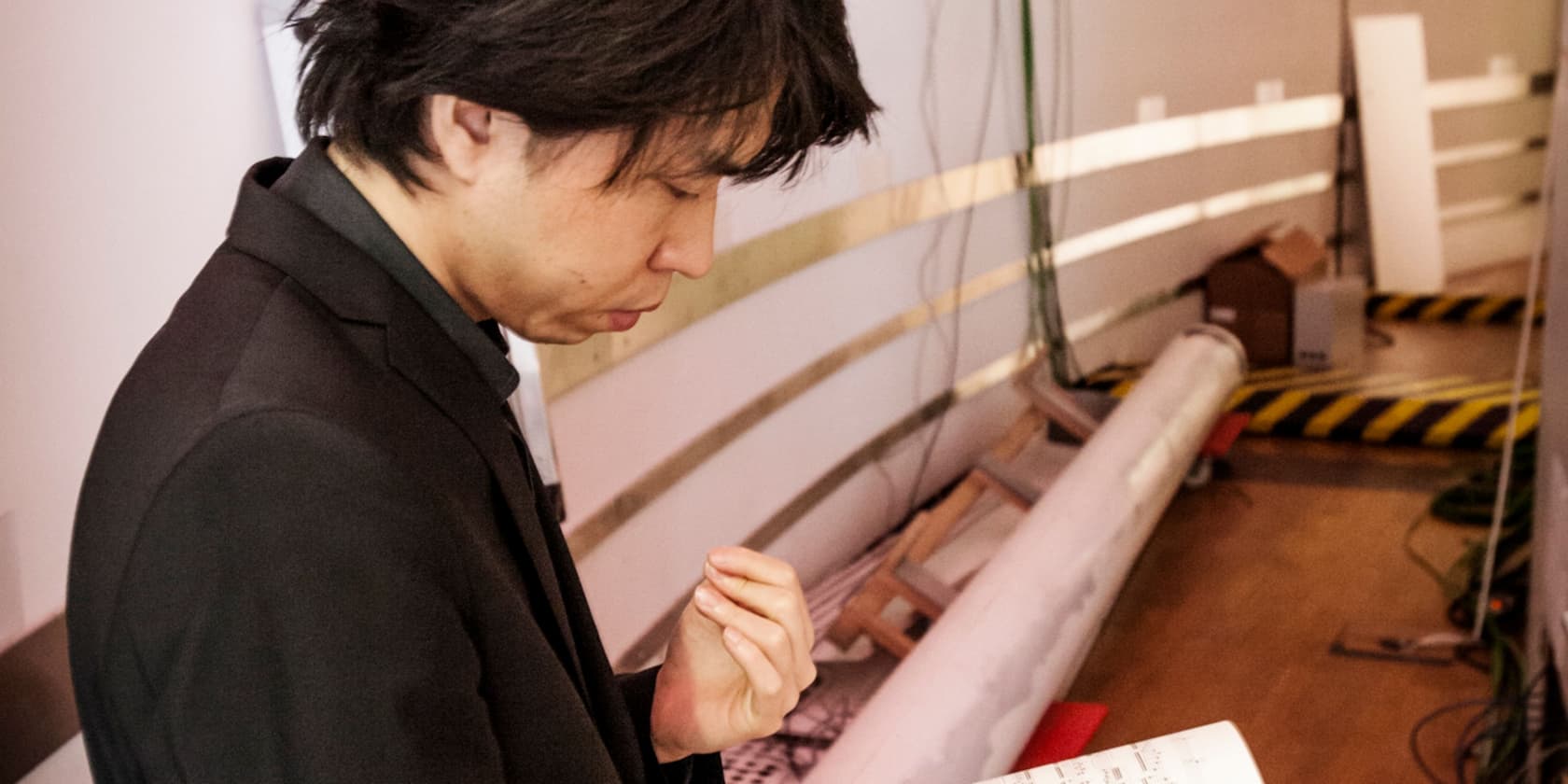 A man in a black suit reads sheet music in a room with various technical equipment.