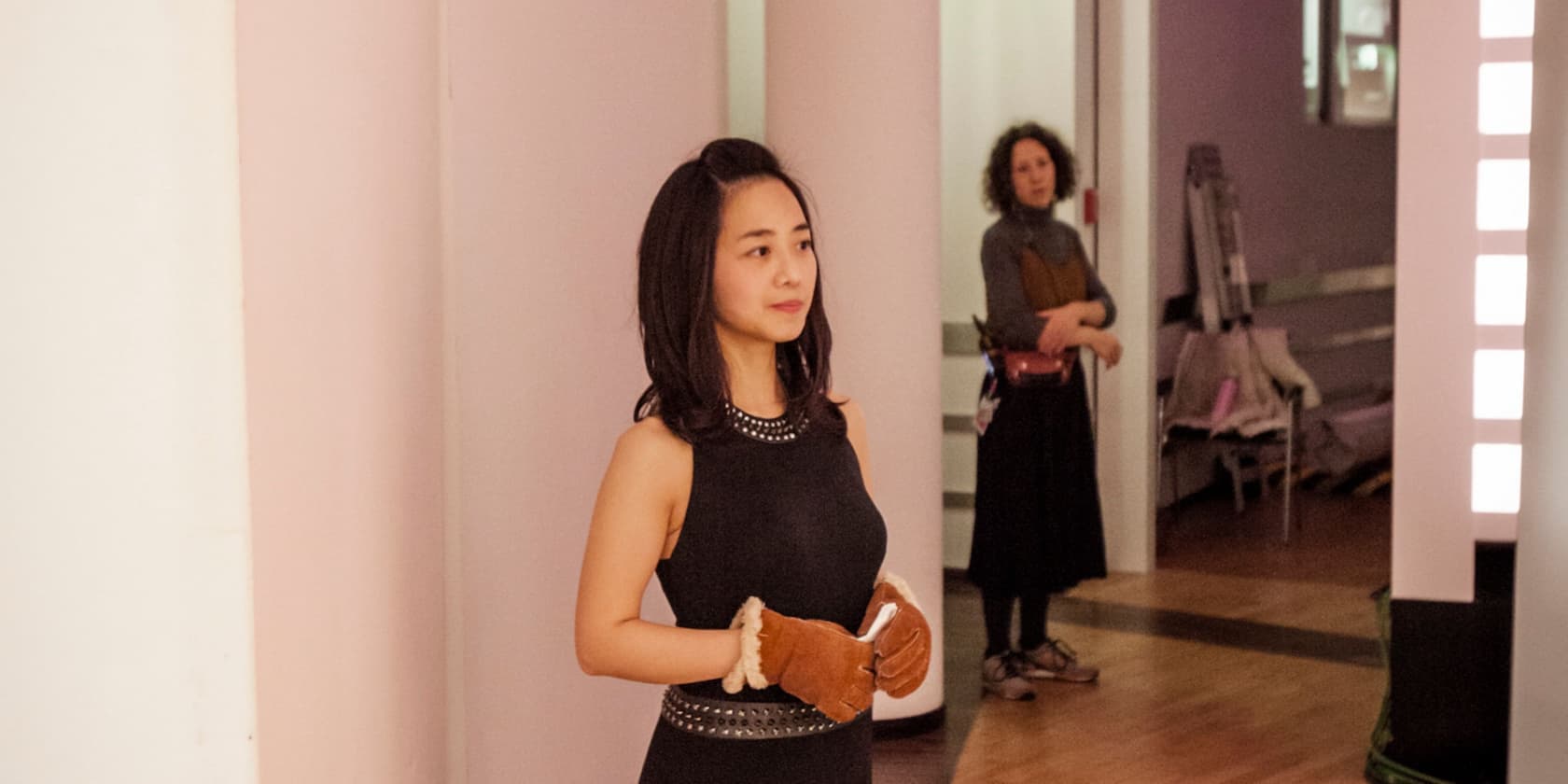 A woman stands in a room wearing brown gloves. Another person can be seen in the background.