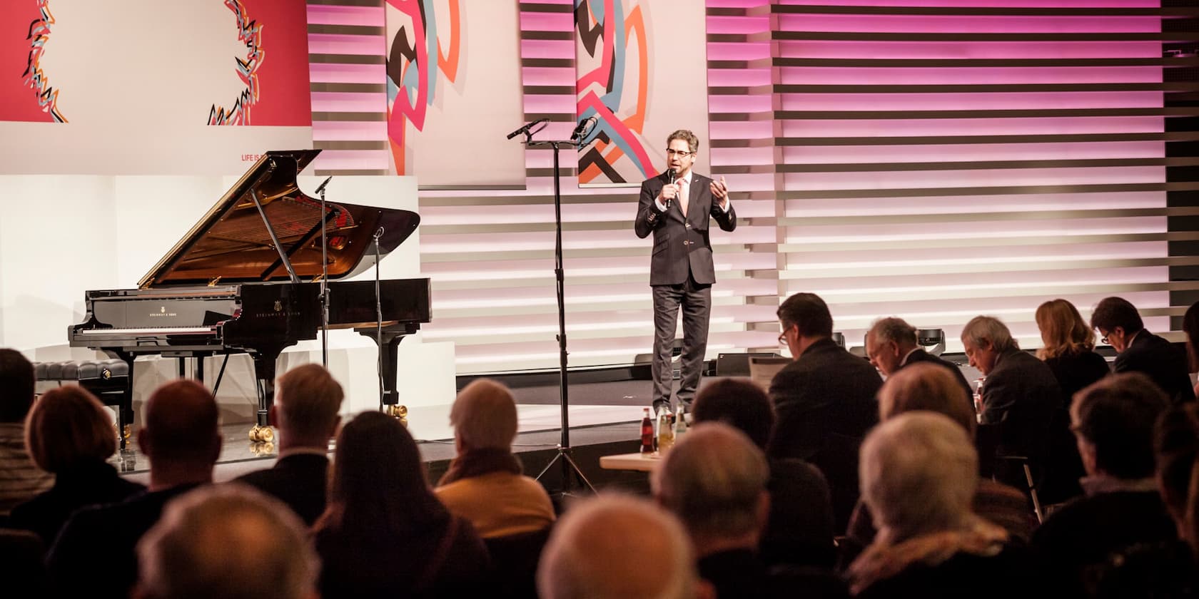 A man gives a speech in front of an audience next to a grand piano on a stage.