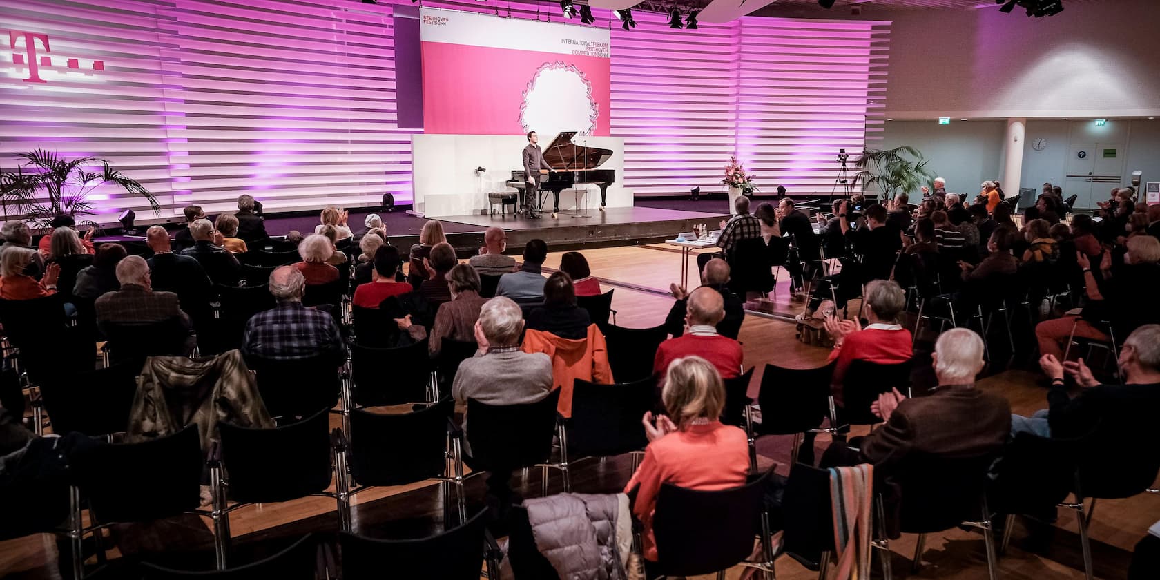 Ein Pianist spielt auf einer Bühne vor einem sitzenden Publikum. Im Hintergrund ist ein großes Logo von Telekom, sowie ein Banner der International Telekom Beethoven Competition Bonn zu sehen.