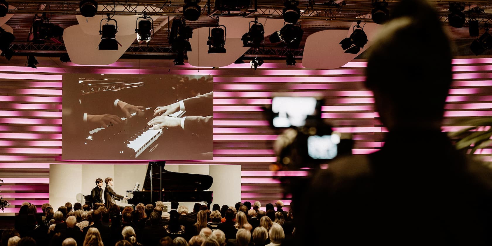 A piano concert with two musicians, while an audience member records with a camera. On stage, a large screen displays close-ups of hands playing the piano.