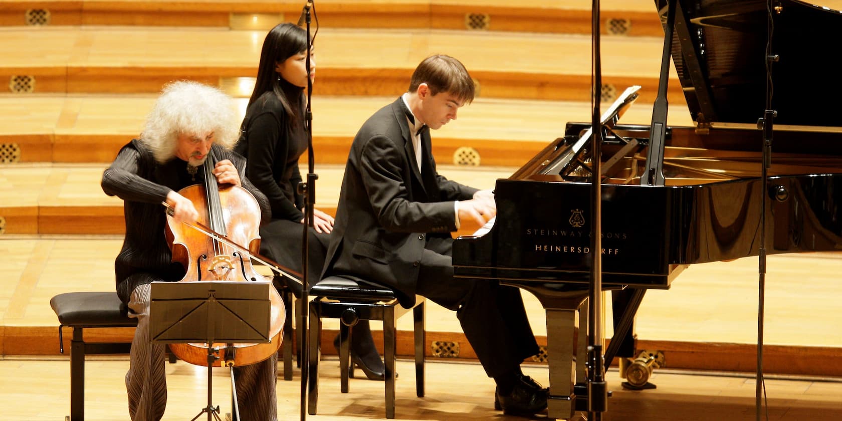 Three musicians at a concert: a cellist playing the cello, a pianist playing the piano, and a third person sitting in the background.