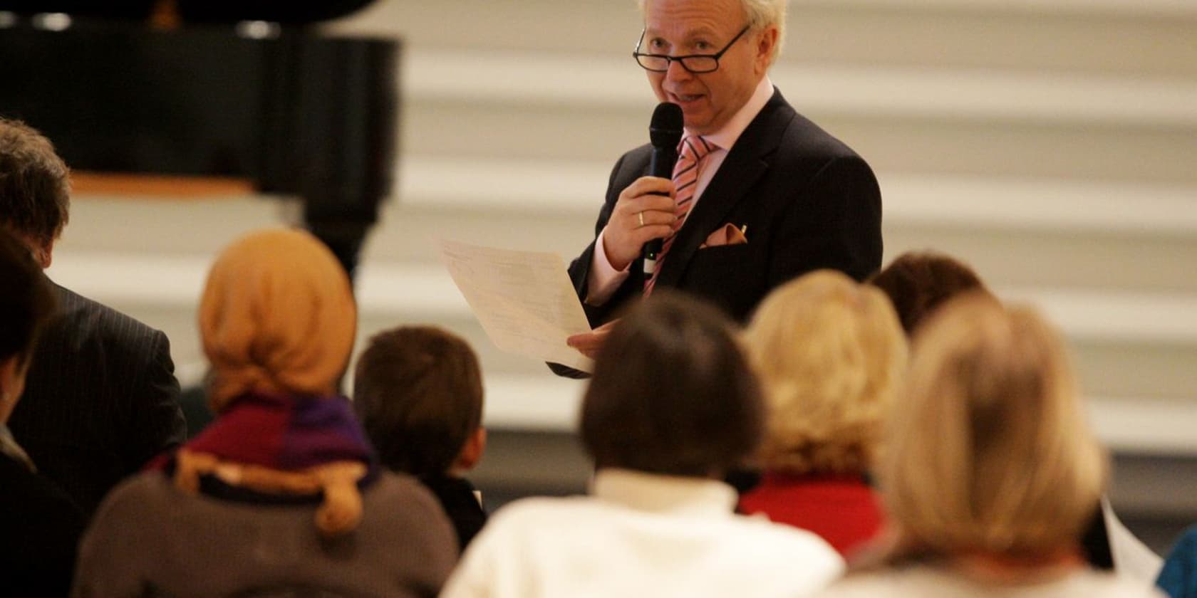 A man holds a microphone and speaks to an audience seated on chairs.