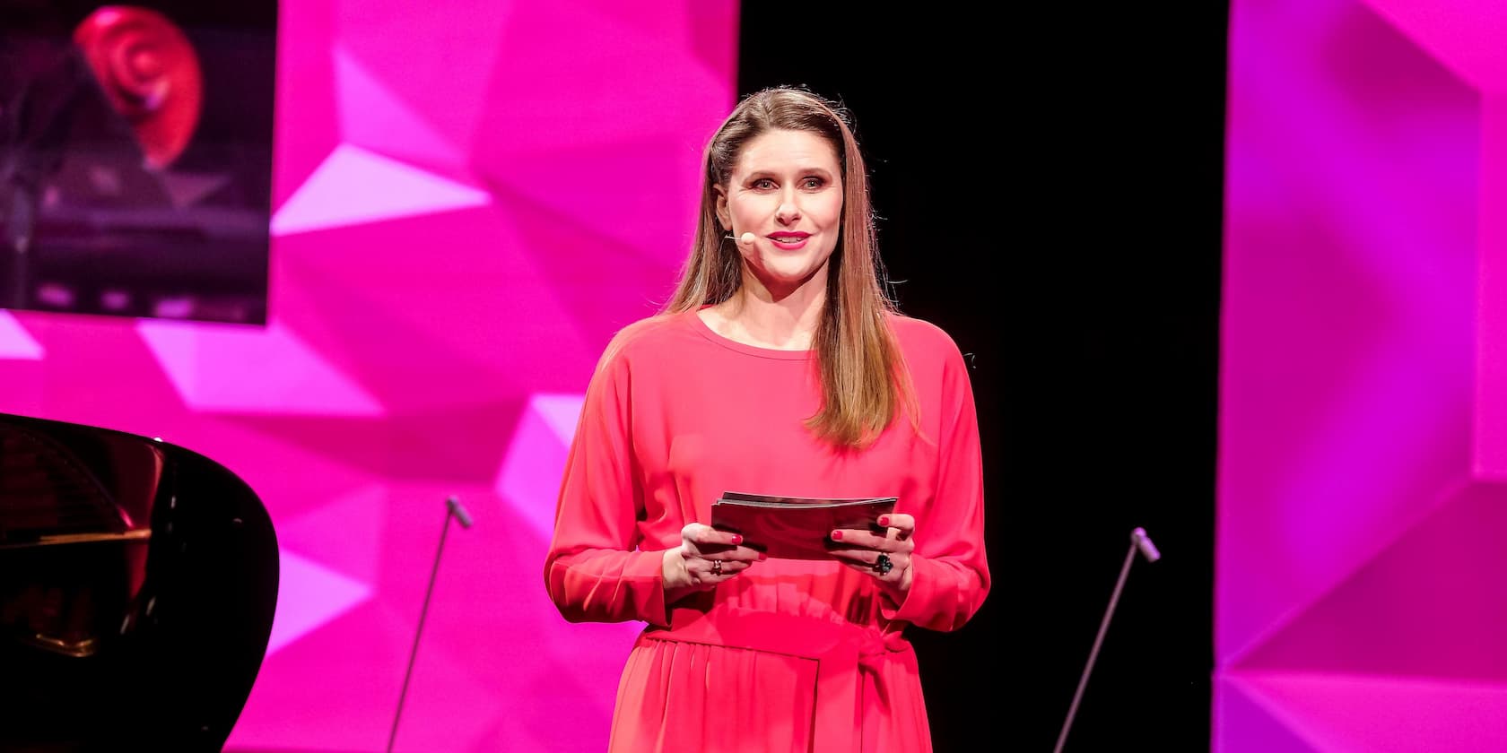 A woman in a bright red dress speaks on a stage with a pink background.