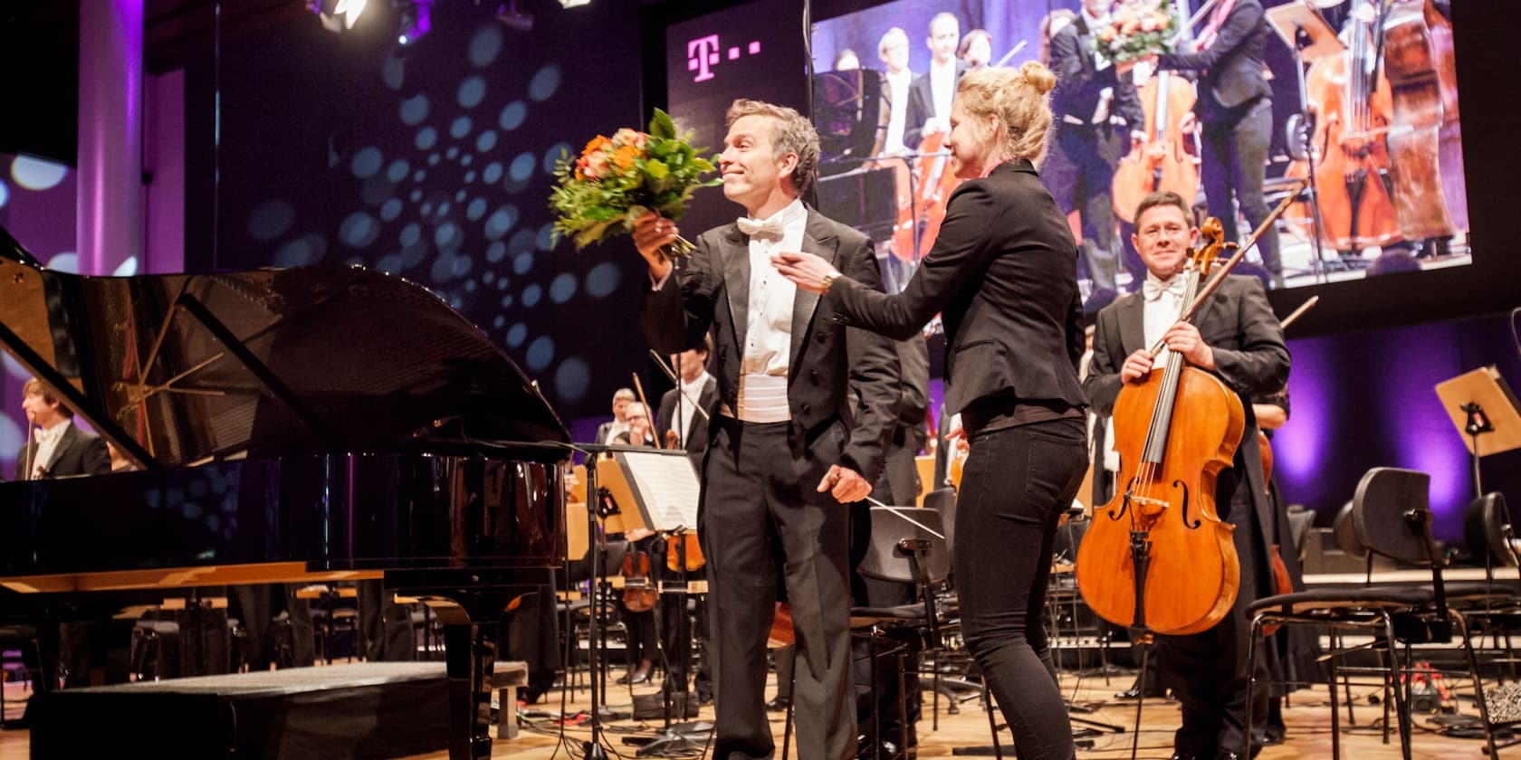 A conductor is being honored with a bouquet of flowers by a woman on stage. Musicians with instruments in the background.