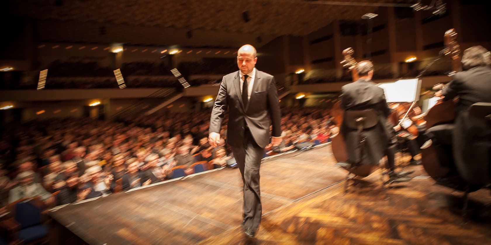 A man in a suit walks across a stage while an orchestra plays in the background. The audience is blurred.