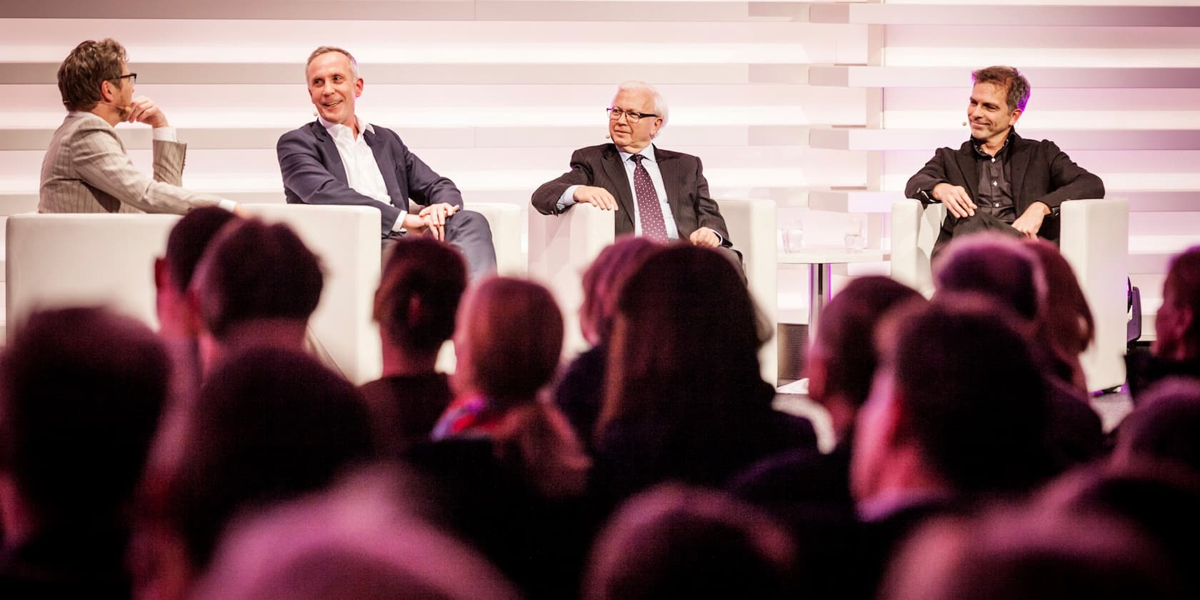 Vier Männer sitzen auf einer Bühne und nehmen an einer Podiumsdiskussion teil, während das Publikum zuschaut.