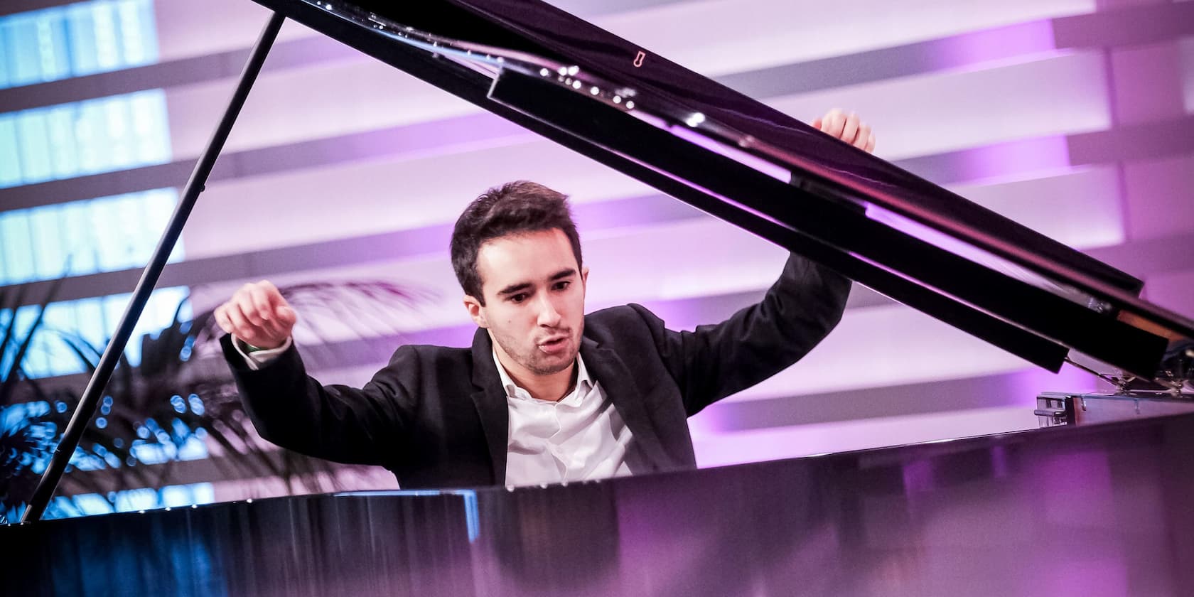 A man energetically plays the piano on a stage with purple lighting.