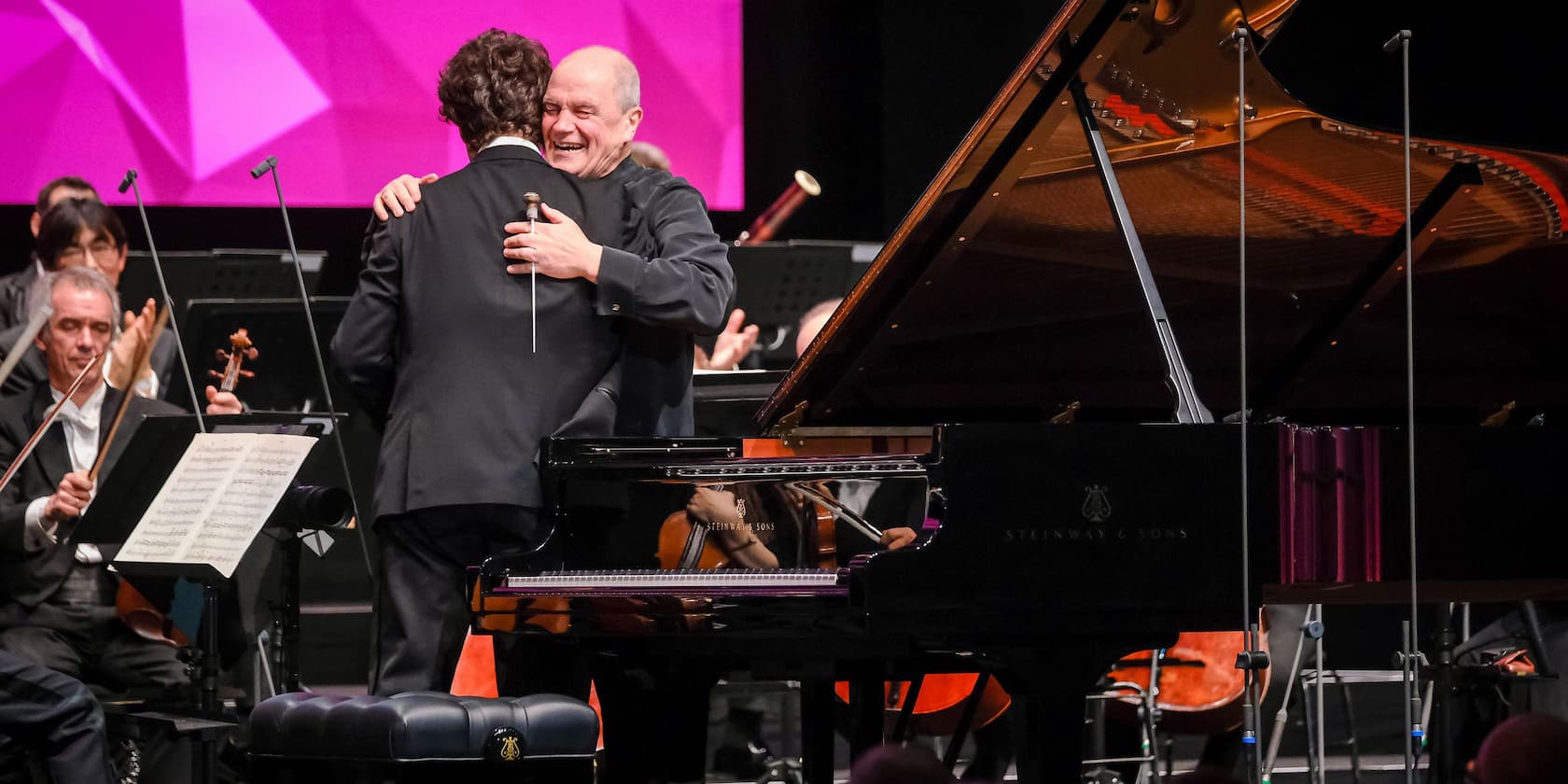 Two men hug on stage after a piano concert. An orchestra in the background applauds.