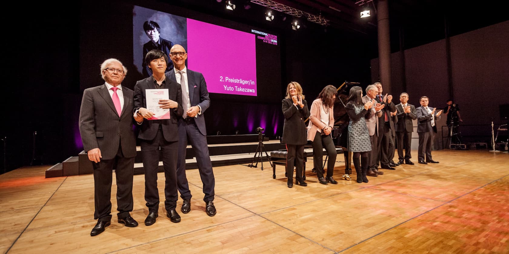 Yuto Takezawa receives the second prize at a competition and stands on stage with other awardees. A large screen in the background shows his name and a photo of him.