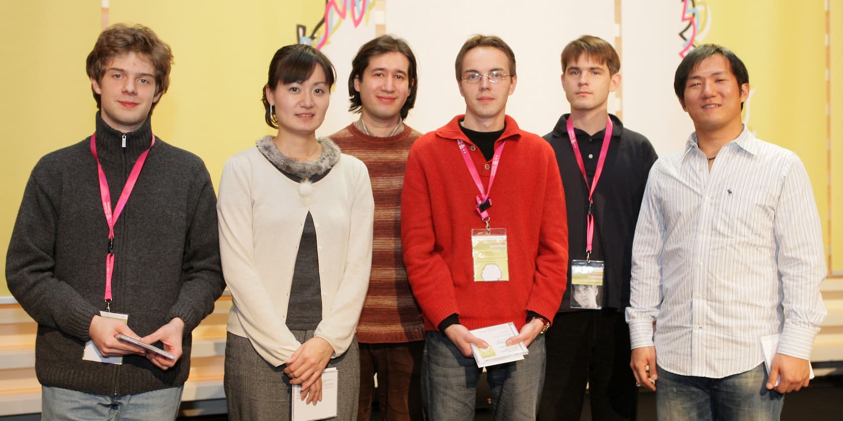 Six young people standing in a row and smiling at the camera. They are wearing different colored clothes and some have name tags hanging around their necks.