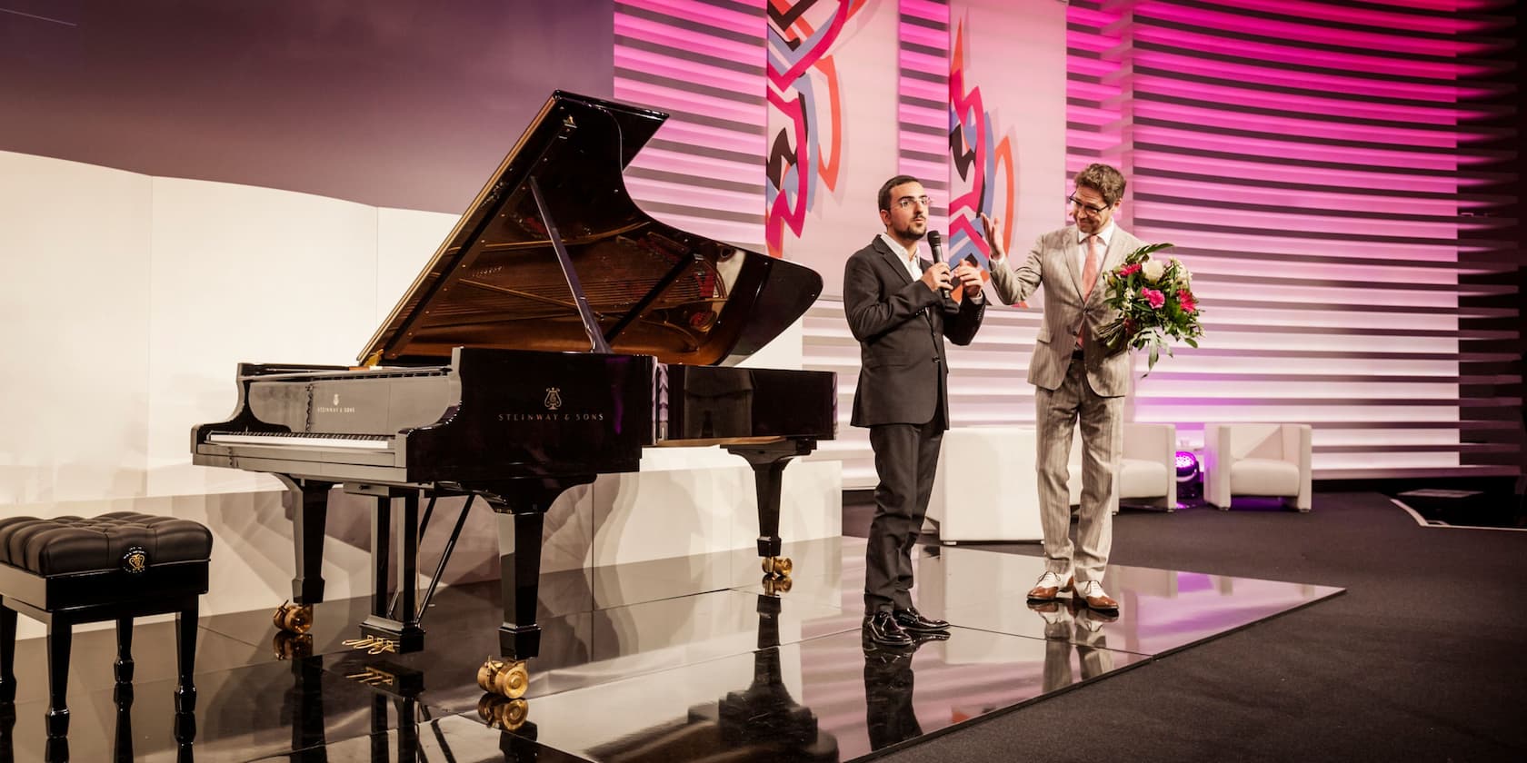 A man is giving a speech with a microphone while another man stands beside him holding a bouquet of flowers. A grand piano and stool are on the left side of the image.