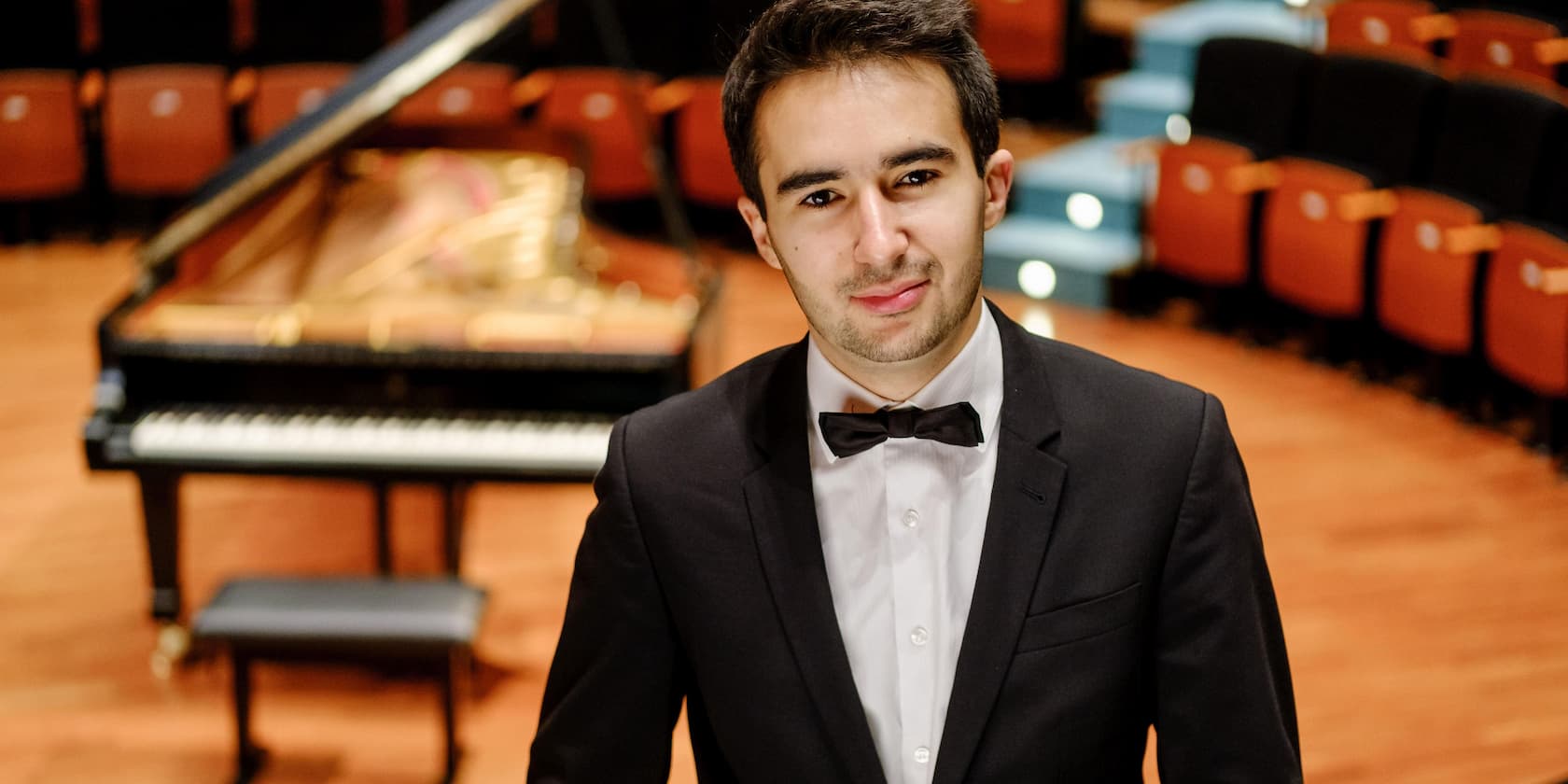 A man in a suit and bow tie stands in front of a grand piano in a concert hall.