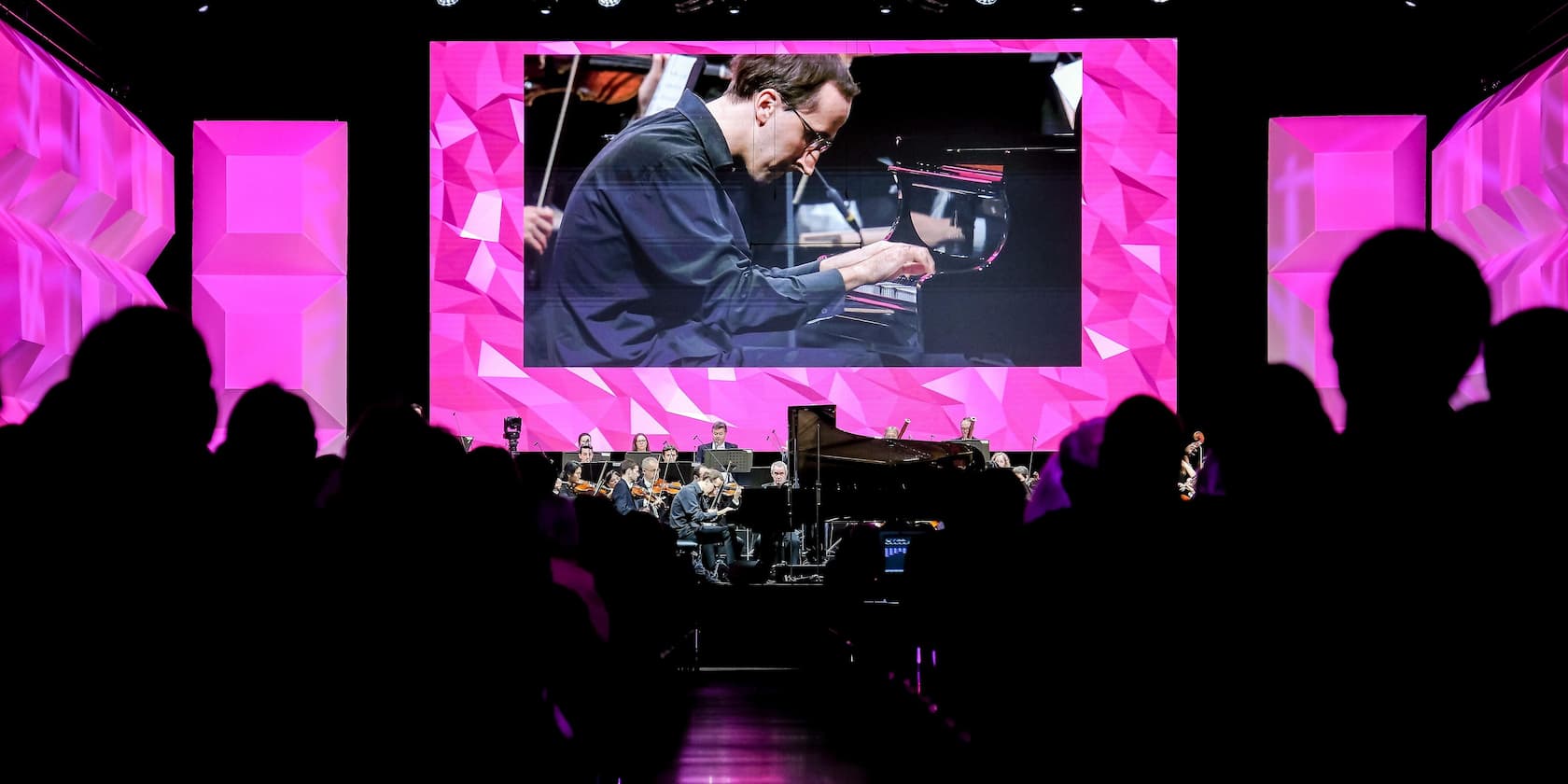 Ein Konzert mit einem großen Bildschirm im Hintergrund, der einen Musiker am Klavier zeigt. Im Vordergrund sind die Silhouetten des Publikums zu sehen.