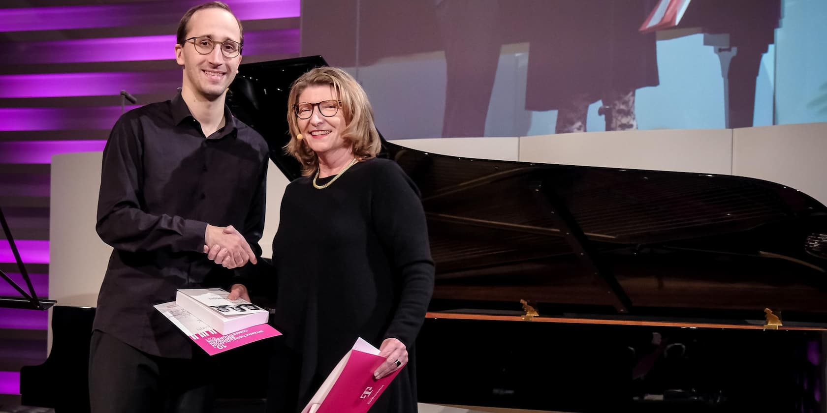 A man holding a certificate and a book shakes hands with a woman in a black dress. A grand piano is in the background.