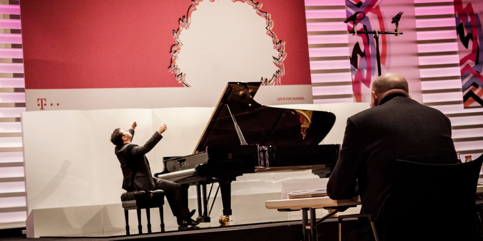 A pianist plays a large black piano on a stage with an audience member in the foreground.