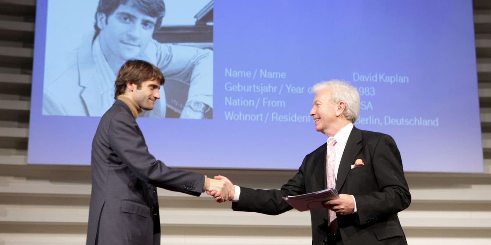 Two men shaking hands on a stage. In the background, a projection screen with the name and details of David Kaplan is visible.