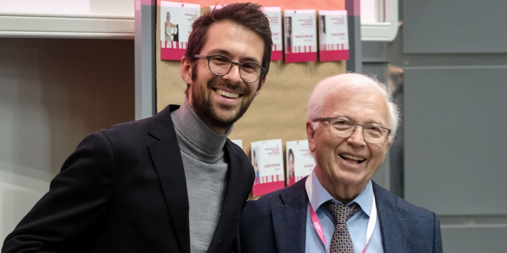 Two smiling men in suits stand in front of a wall with multiple ID cards.