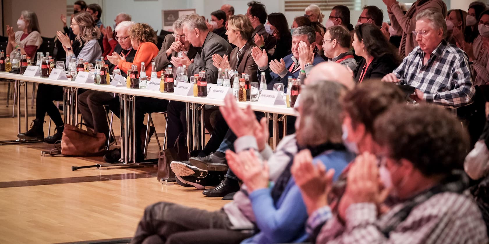 An audience applauds in a large hall. Drinks and nameplates are on the table in front of them.