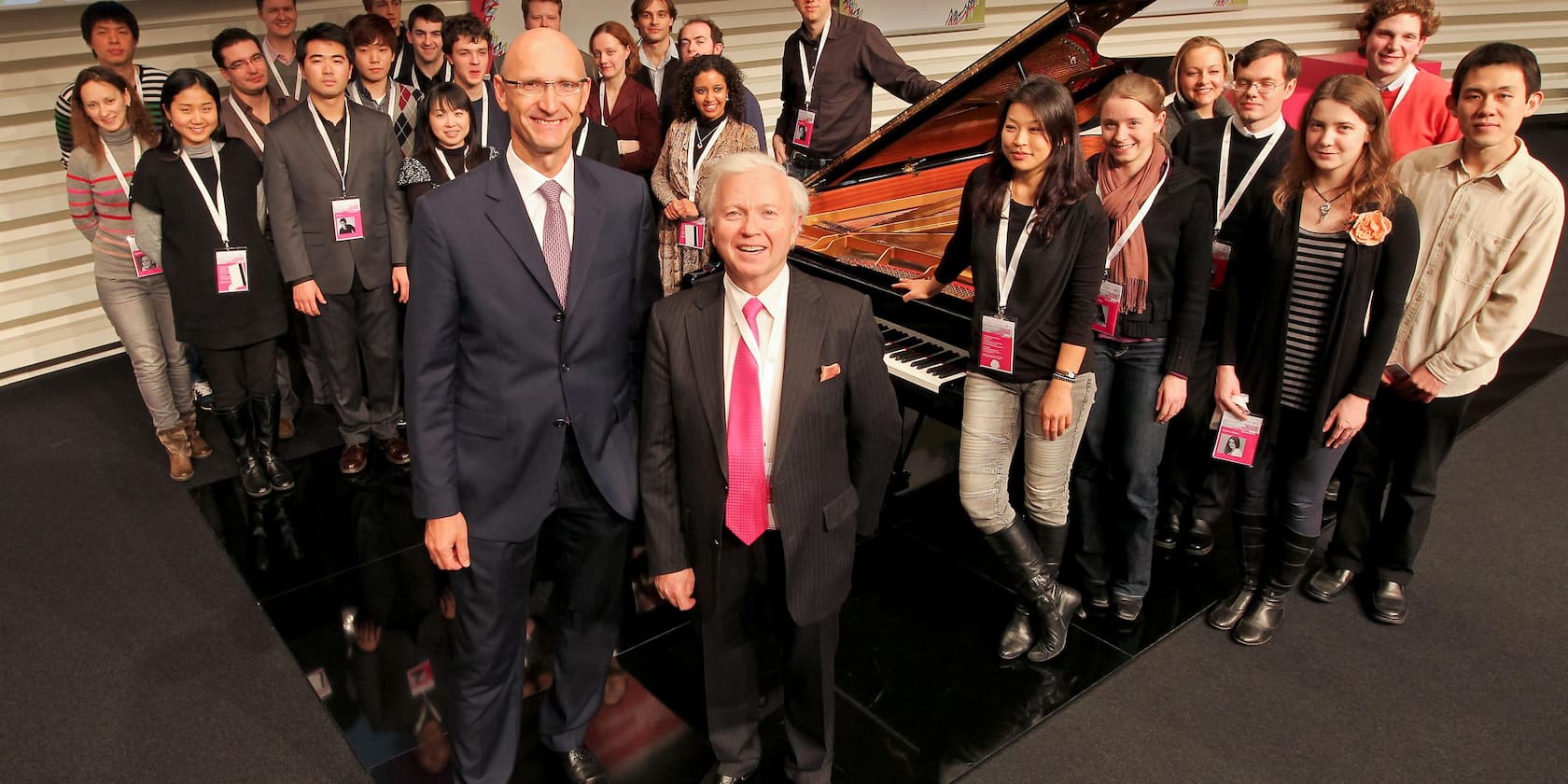 Group of people standing in front of a grand piano, two men in the foreground.