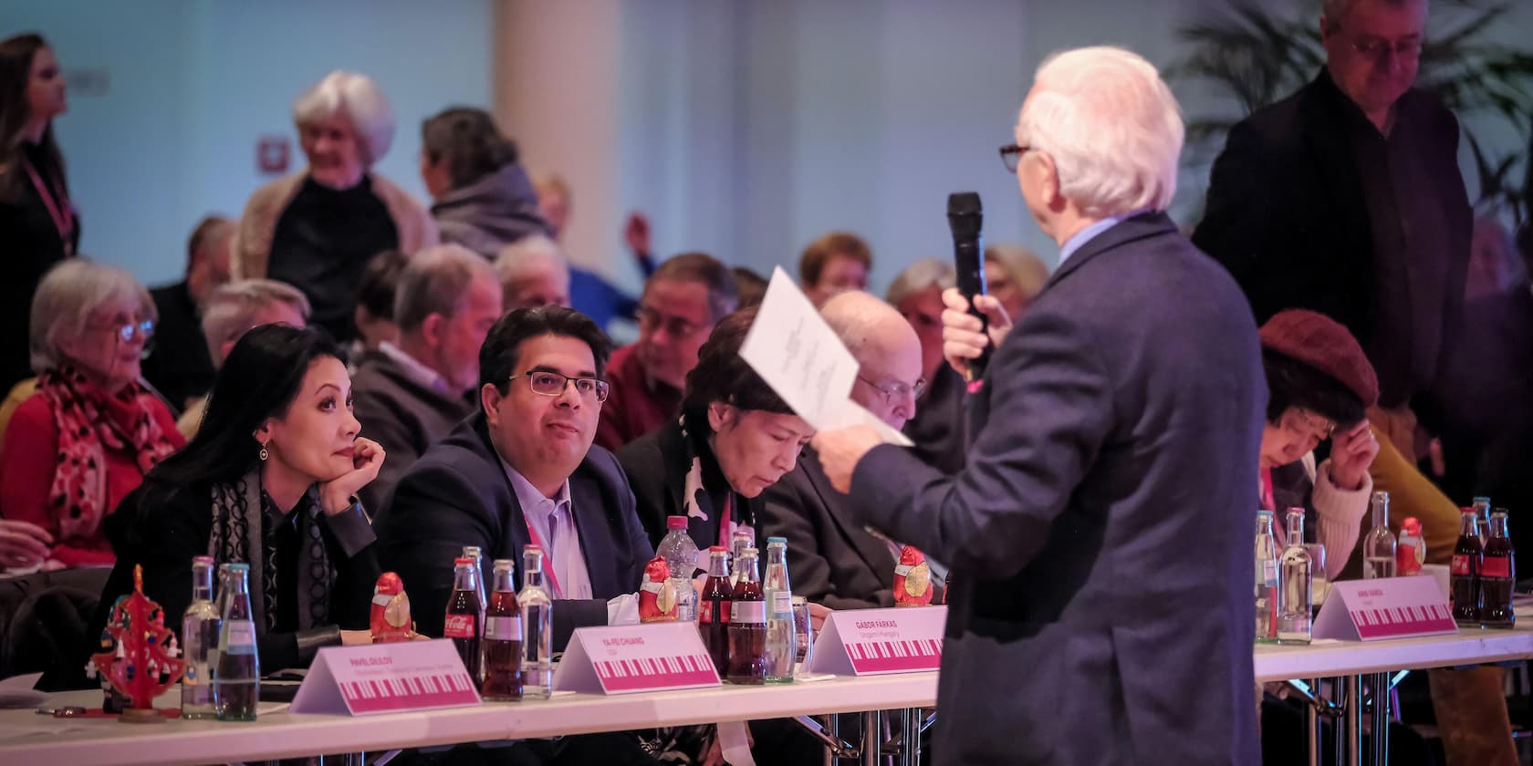 A man speaks with a microphone in front of a group of people sitting at a table covered with drinks.