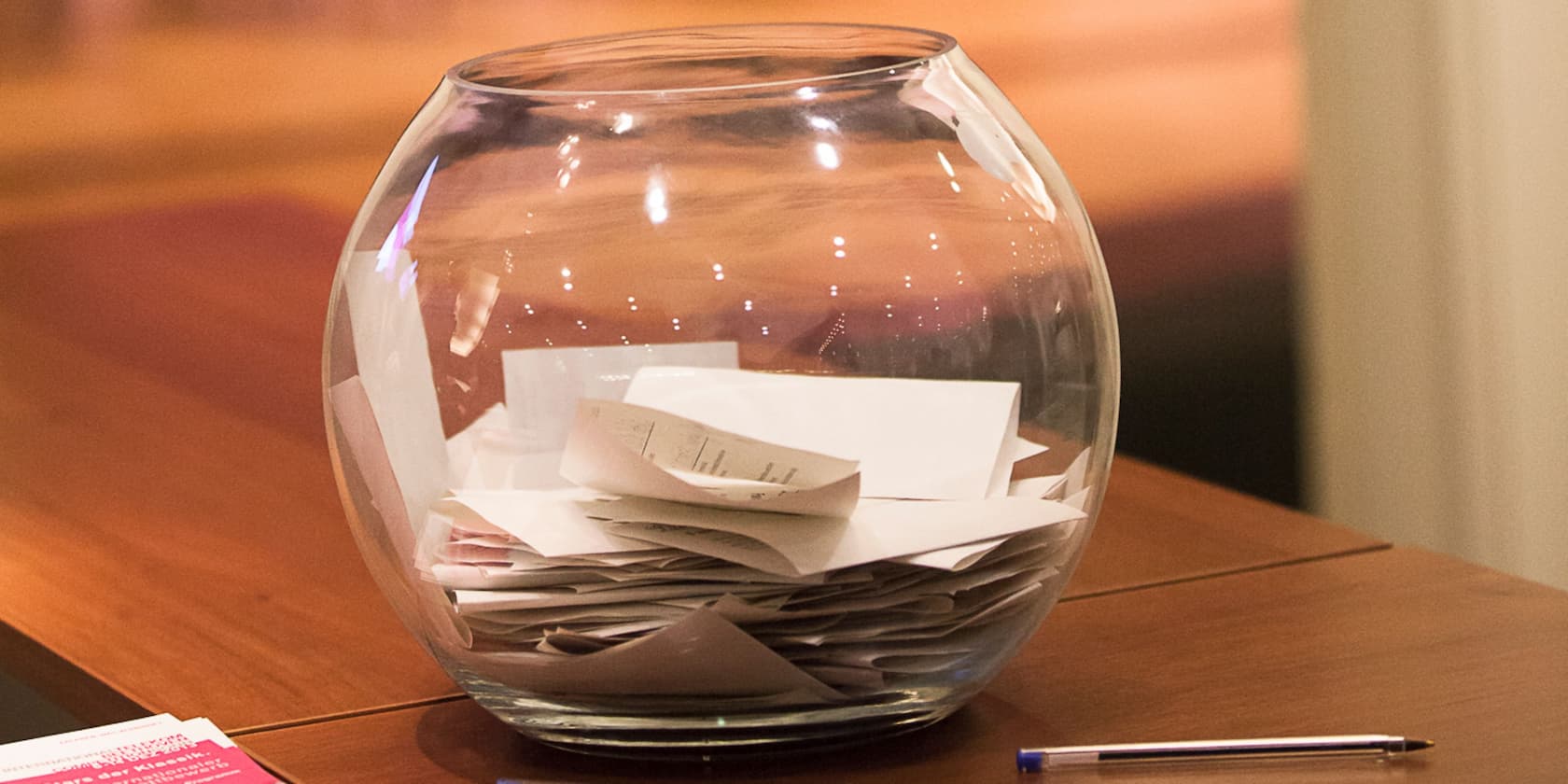 A glass container filled with folded slips of paper sits on a table. A pen lies next to it.